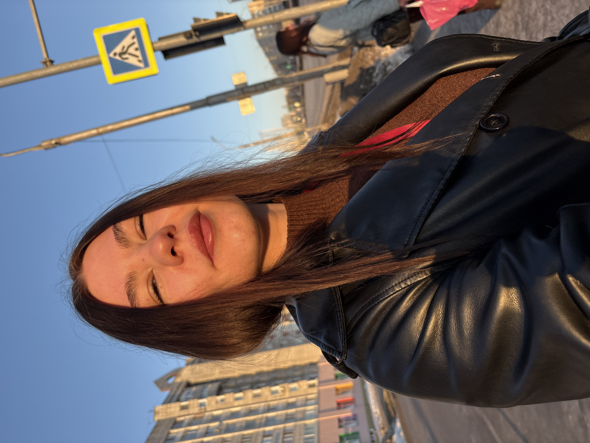 A low-angle shot captures a woman with long brown hair, a brown sweater, and a black leather jacket against an urban backdrop. Her eyes are closed, and she has a slight smile. In the background, a pedestrian crossing sign is visible against a clear blue sky. A city building with many windows is situated behind her. Given the presence of a pedestrian crossing sign and the architecture of the building, it's plausible this photo was taken in Perm, Russia. Other people are visible in the blurred background.
