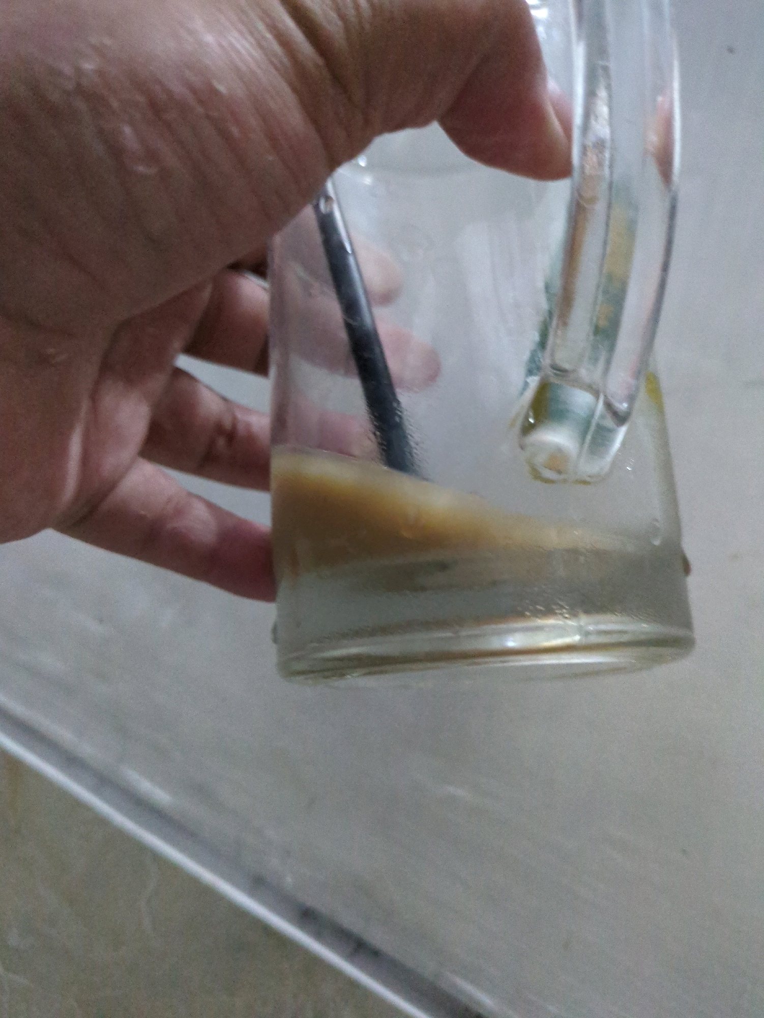 A person's hand is holding a clear glass mug with a handle. Inside the mug, there is a small amount of liquid at the bottom, with a layer of sediment or residue on top of it. A dark-colored straw is partially submerged in the liquid. The mug is resting on a textured surface, possibly a countertop, which appears to be a sink area. The lighting is dim, and the focus is primarily on the mug and the hand holding it. The background is blurred and indistinct. There is no visible indication of the specific location within Sindon, Indonesia.