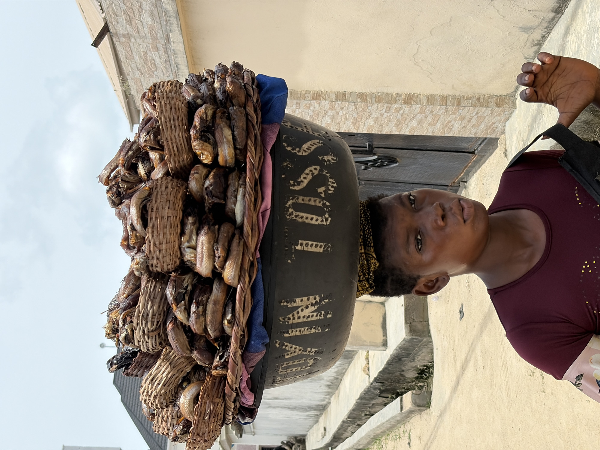 An outdoor, eye-level portrait-style photograph depicts a person carrying a substantial load of dried fish on their head, likely a vendor in Igboefon, Nigeria. The image is taken from a slightly low angle, emphasizing the towering load.
The primary subject is a person, likely a woman, with dark skin and a neutral expression, looking directly at the camera. She wears a maroon short-sleeved top adorned with small golden embellishments on the shoulder, and a floral-patterned garment (skirt or lower half of a dress) is visible on her right side. Her dark hair is styled, and a patterned headwrap or band, in yellow and black, is visible beneath the heavy load.
Balanced on her head is a very large, dark, circular container, possibly a basin or calabash, which serves as a base. On top of this container, a massive stack of dried or smoked fish is meticulously arranged within numerous small, woven baskets and bundles, creating a tall, conical shape. A blue and a pink cloth are tucked between the fish baskets and the large container, possibly for cushioning or decoration.
Visible text on the side of the large dark container reads "MAI YAM USSD" in white or light yellow, stenciled, partially worn letters. "MAI YAM" is clearly discernible, followed by "USSD," which is a common mobile payment protocol in Nigeria.
The background shows an outdoor setting with a light-colored building facade, featuring a section of light-brown textured stone cladding. The ground appears to be concrete or paved, with some sparse green vegetation visible. The sky is bright but overcast or partly cloudy, indicating daytime. The scene captures a common method of goods transportation and hawking in the region, reflecting daily life and commerce.
