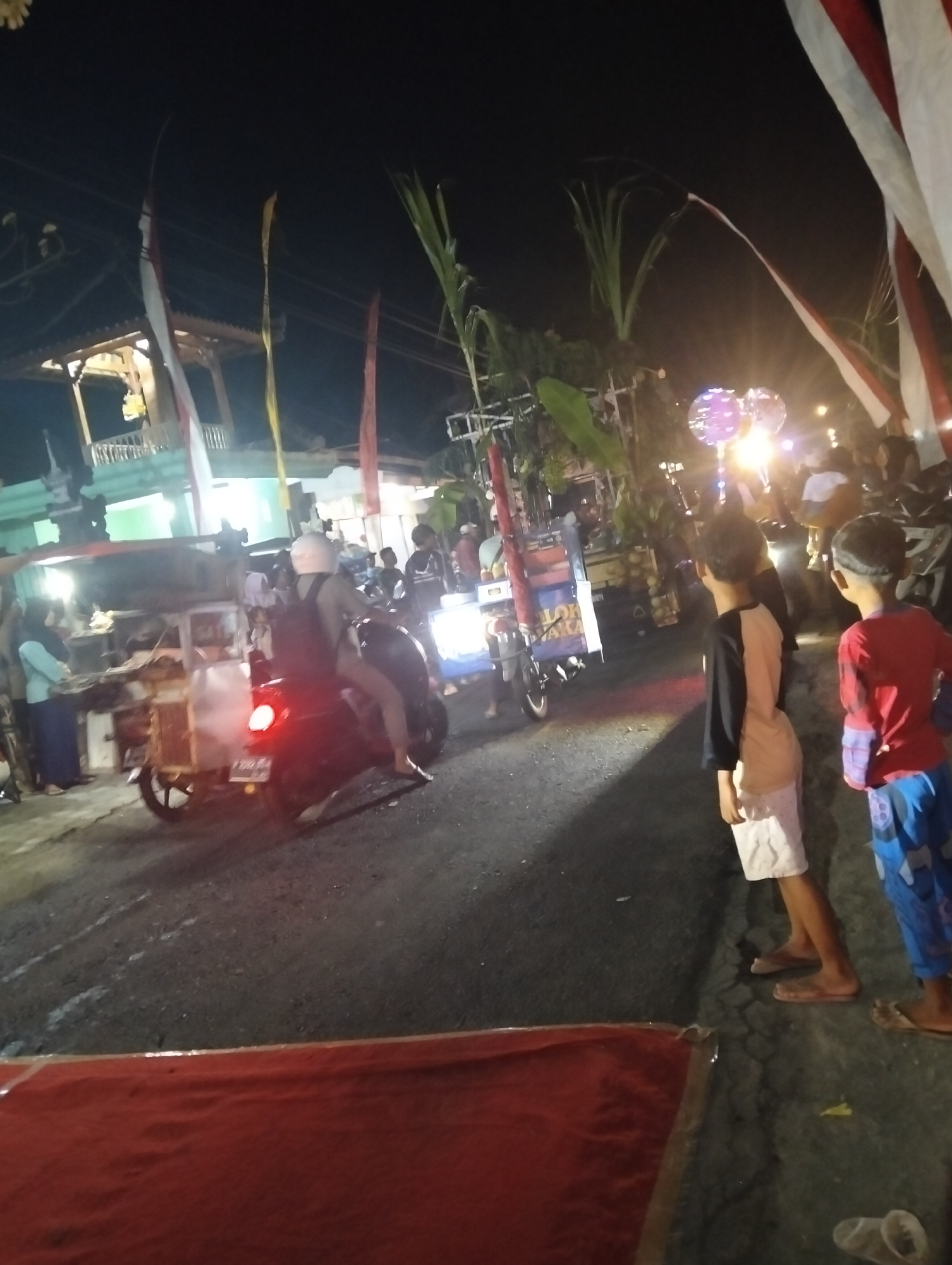 An outdoor nighttime scene in Sumbersewu, Indonesia, depicts a street filled with people and vehicles. A person wearing a white helmet is riding a dark-colored motorcycle. To the left, a food cart with a colorful awning is visible, with individuals standing nearby. In the background, there are buildings adorned with flags, and palm fronds extend into the frame. Several other motorcycles and people are scattered throughout the scene, with some individuals in the foreground standing and observing. The ground is asphalt, with some white markings.