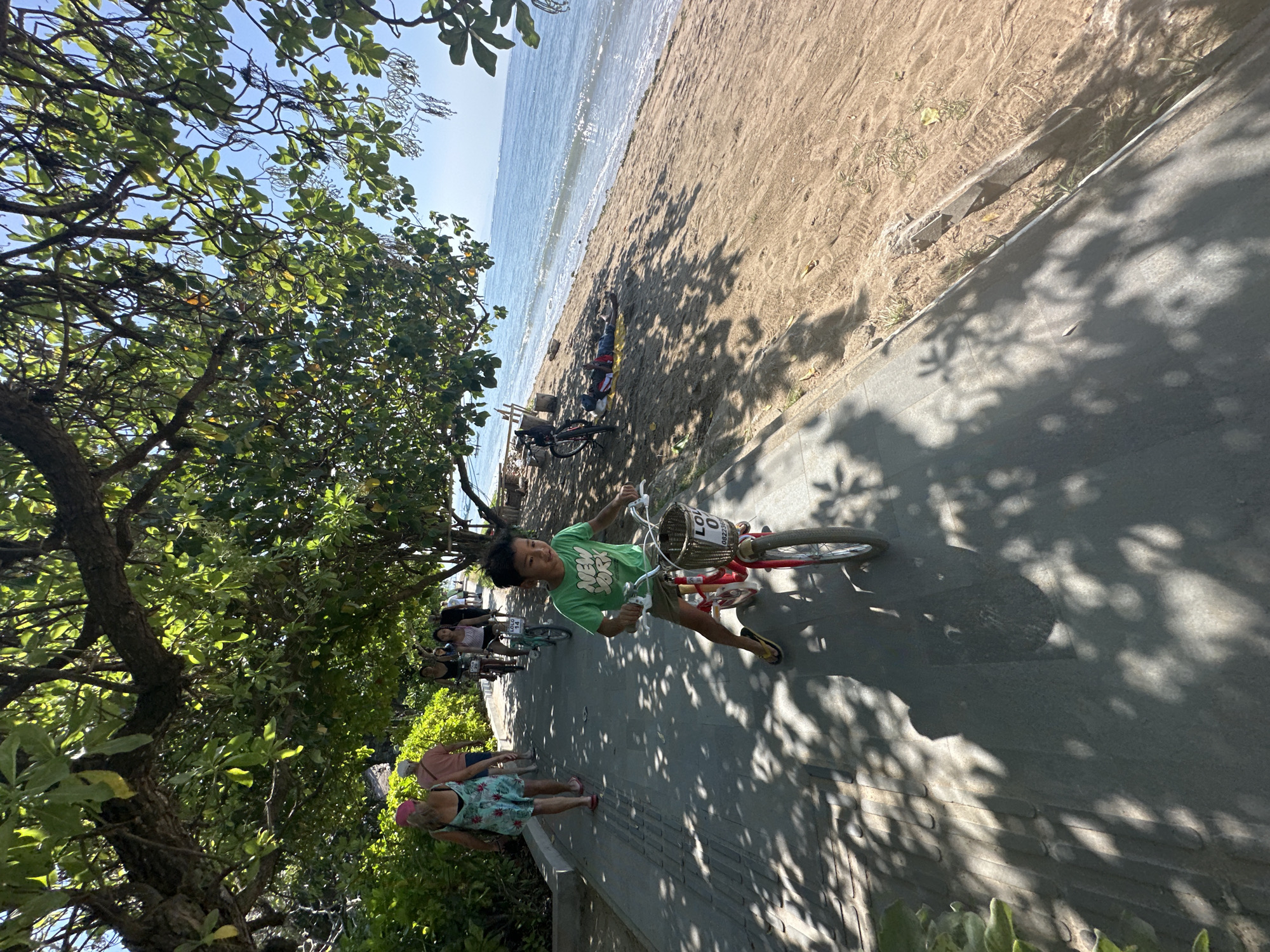 In Denpasar, Indonesia, a young boy rides a red bicycle with a basket on a paved walkway that runs alongside a sandy beach. The boy wears a green t-shirt with "NEW YORK" printed on it and dark shorts, with yellow flip-flops. The walkway is shaded by lush green trees. To the left of the walkway, people are walking and cycling. In the background, the ocean is visible with gentle waves. Further down the beach, a person is lying on a yellow mat under the shade of a tree, with a bicycle nearby. The scene appears to be a sunny day with clear skies.