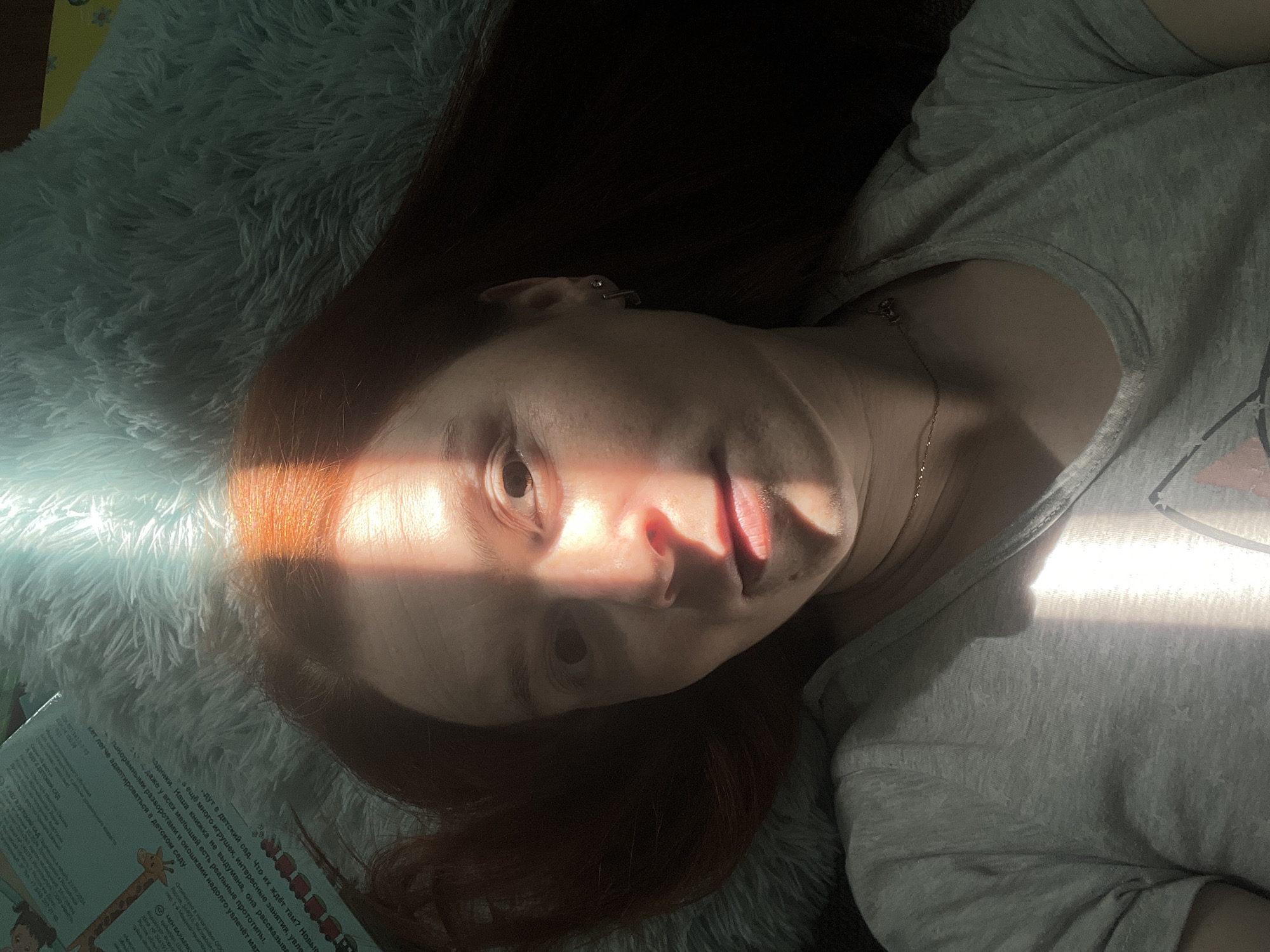 A woman with red hair lies on a fluffy, white surface, possibly a pillow or blanket. She is wearing a grey t-shirt and a gold necklace. A beam of sunlight falls across her face. A children's book is visible in the lower left corner, with text in Russian, giving it a Rostov location within Russia, possibly within Chaikovsky city if specified.
