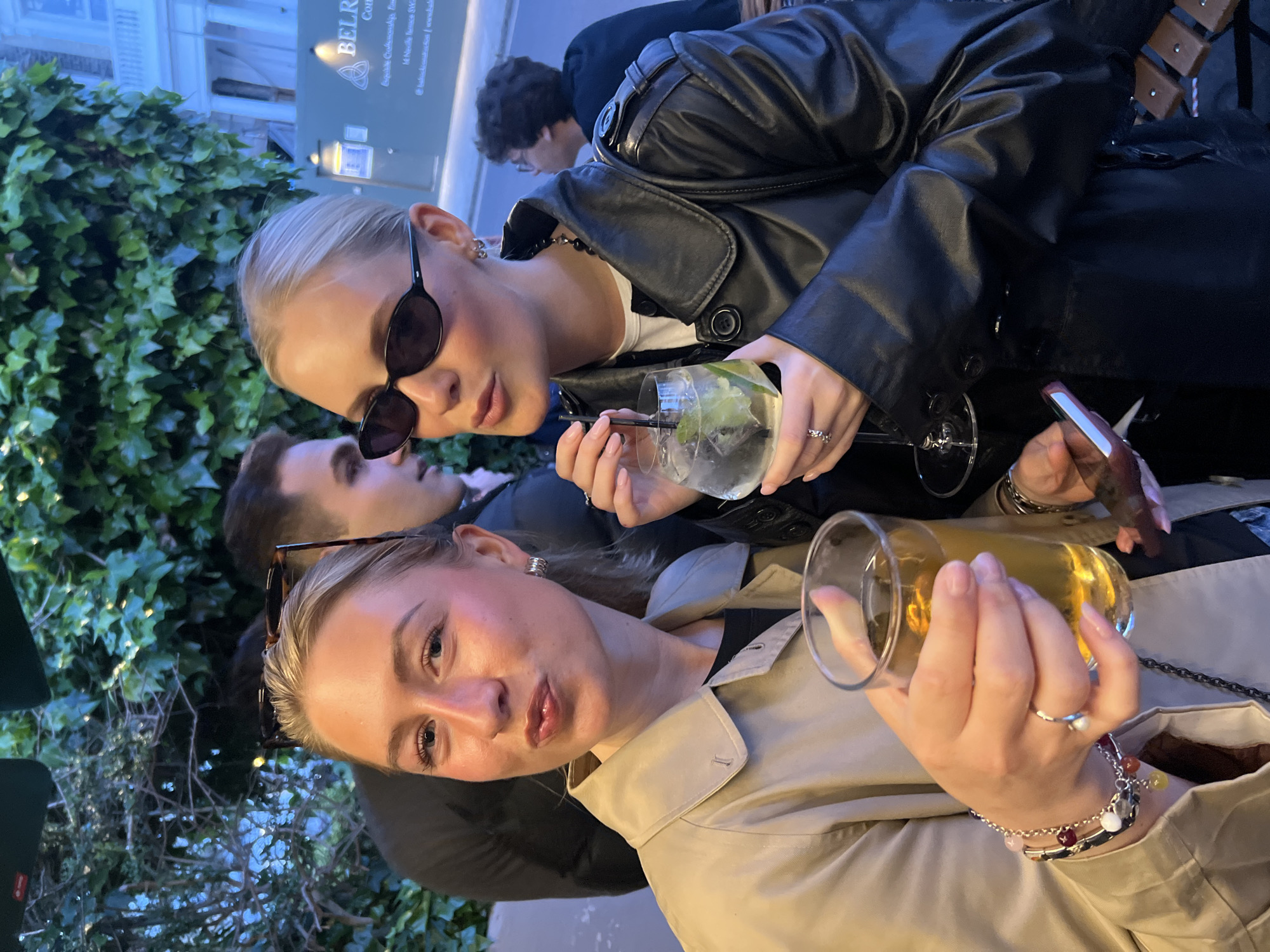 Two young women pose holding drinks, an older man stands behind. The woman in the foreground wears a tan jacket and holds a glass of amber liquid, she wears sunglasses in her hair and a bracelet on her wrist. The woman behind her wears a black leather jacket and black sunglasses, she holds a cocktail glass with a lime. Both women wear rings. A third man stands in the background. The setting appears to be outdoors near a building with climbing ivy and a sign reading "BELR" which could indicate the Belgravia area of London, United Kingdom.
