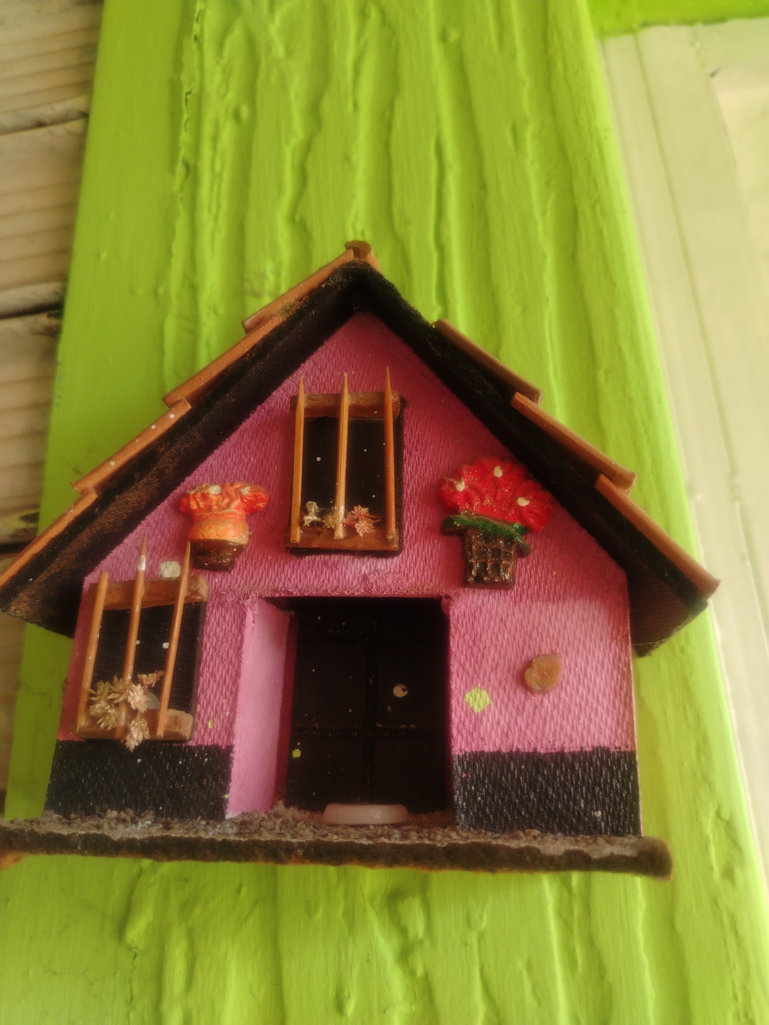 A pink miniature house with a brown roof is attached to a vibrant green wooden surface. The house has a black base and features two small windows with wooden bars and dried floral decorations, and one small, brown, oval-shaped stone. Above the windows, there are decorative red flower pots. A dark brown door is visible in the center. The background consists of a bright green, textured wooden surface and a glimpse of what appears to be a light-colored wall or window frame. The image appears to be a close-up photograph taken at a slightly angled perspective. The location is in the city of Juan Griego, Venezuela.
