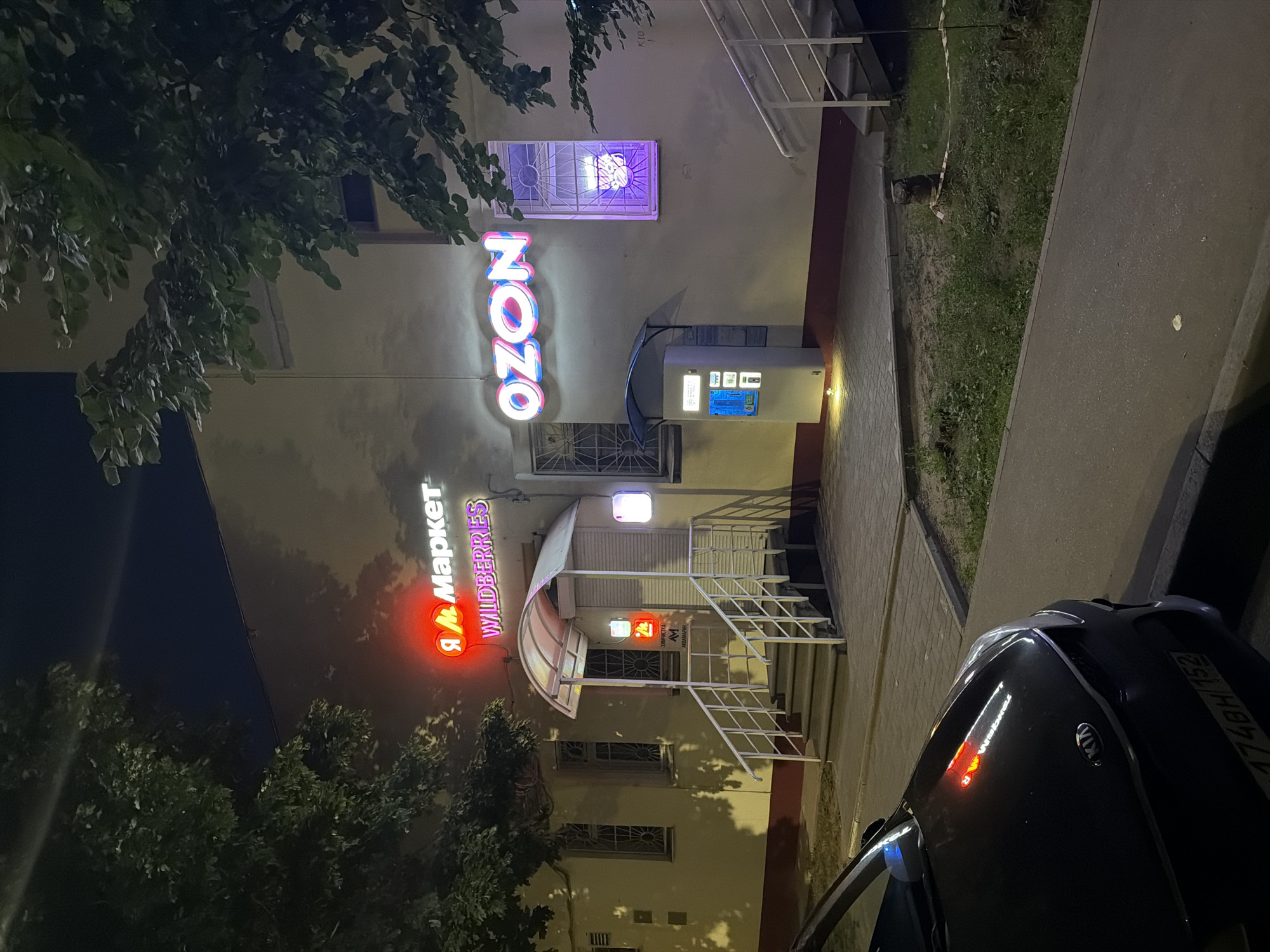 The image shows a storefront at night in Nizhny Novgorod, Russia. A red neon sign that reads "Я Маркет" (Ya Market) with "WILDBERRIES" below it is visible on the building. To the right, a blue and pink neon sign reads "OZON". There is a white kiosk with a blue screen in front of the OZON sign. A dark-colored Kia car is parked in front of the building. The building has a light-colored facade with several windows and a covered entrance with stairs. Trees with green leaves are visible on the left and right sides of the image. The ground in front of the building is paved, with grass and some dirt patches.