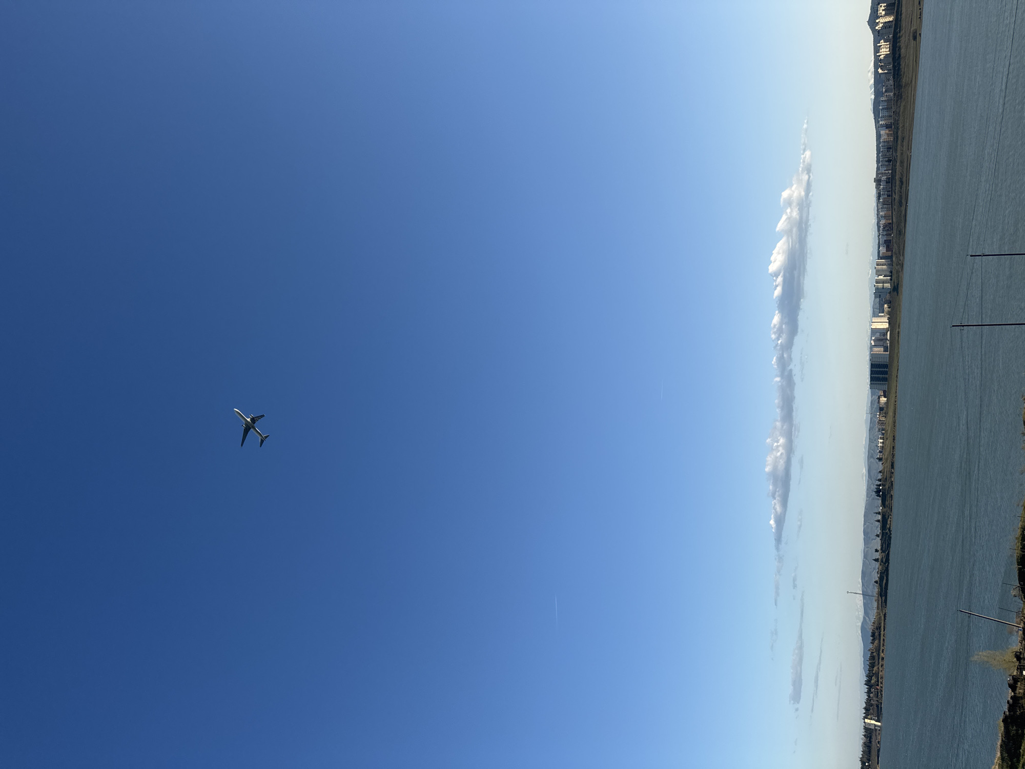 The image shows a split view of a clear sky and a landscape. The left side of the image is predominantly a deep blue sky with an airplane flying in the distance. A faint contrail can be observed in the sky as well. The right side reveals a landscape consisting of a body of water, possibly a river or a lake, running alongside a stretch of land. Along the land, there are urban structures, including buildings and what appears to be city skylines, as well as trees. In the sky near the edge of the water is a long, vertical cloud formation. The image is rotated 90 degrees clockwise. While the information is not present in the image, the request specifies that this image is taken in Tbilisi, Georgia.