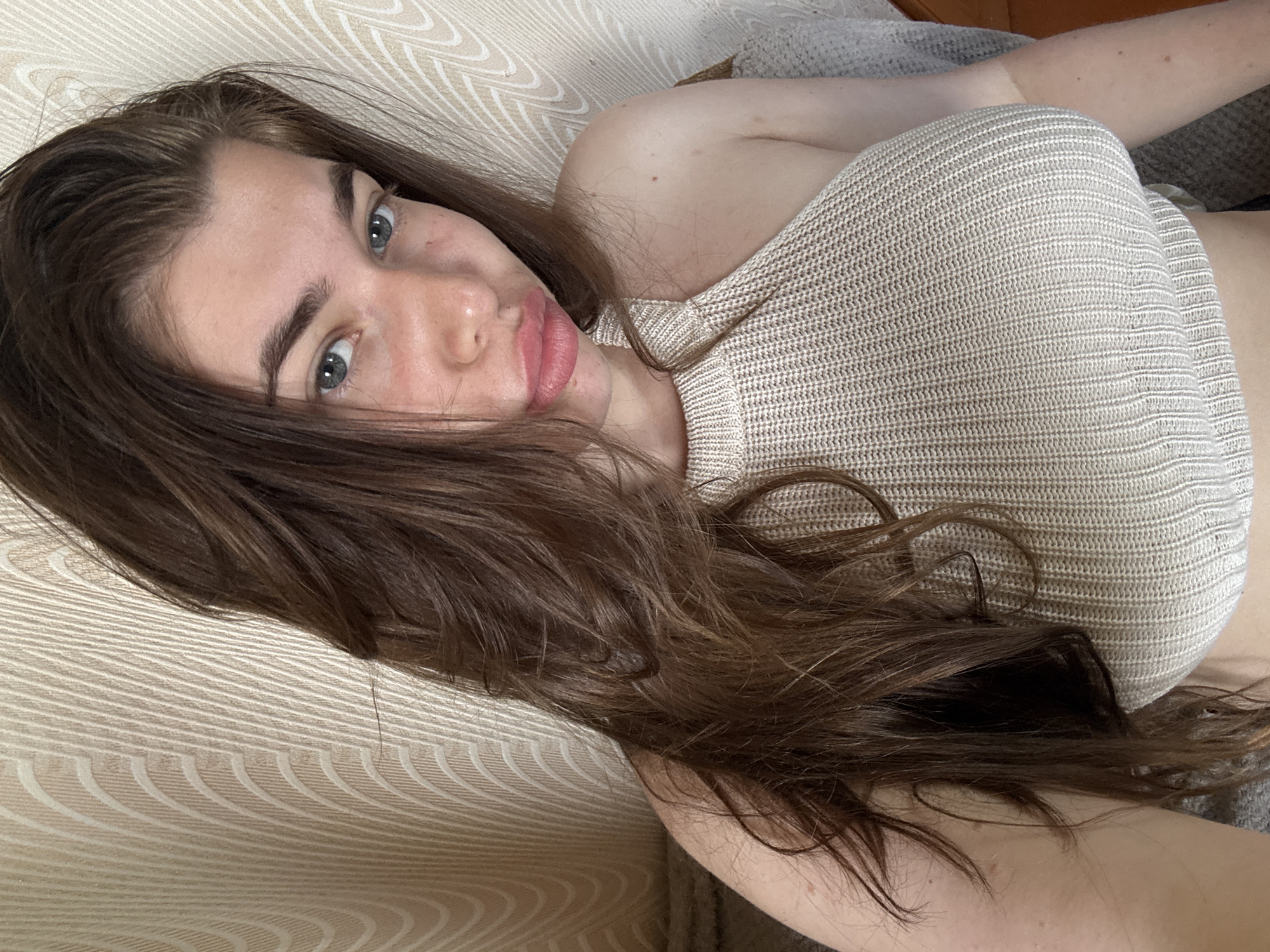 This media file shows a close-up, horizontally oriented shot of a young woman with long, dark brown hair, lying down and looking at the camera. She is wearing a light-colored, knitted top. Her hair is spread out around her, partially obscuring her body. She has fair skin and light blue eyes. Her gaze is direct, and her expression appears relaxed. The background is a textured surface, possibly a sofa or couch, with a beige and white patterned fabric. The lighting suggests it is daytime, with soft natural light. There are no visible text elements in the image. The location context provided is Мещёры, Russia.