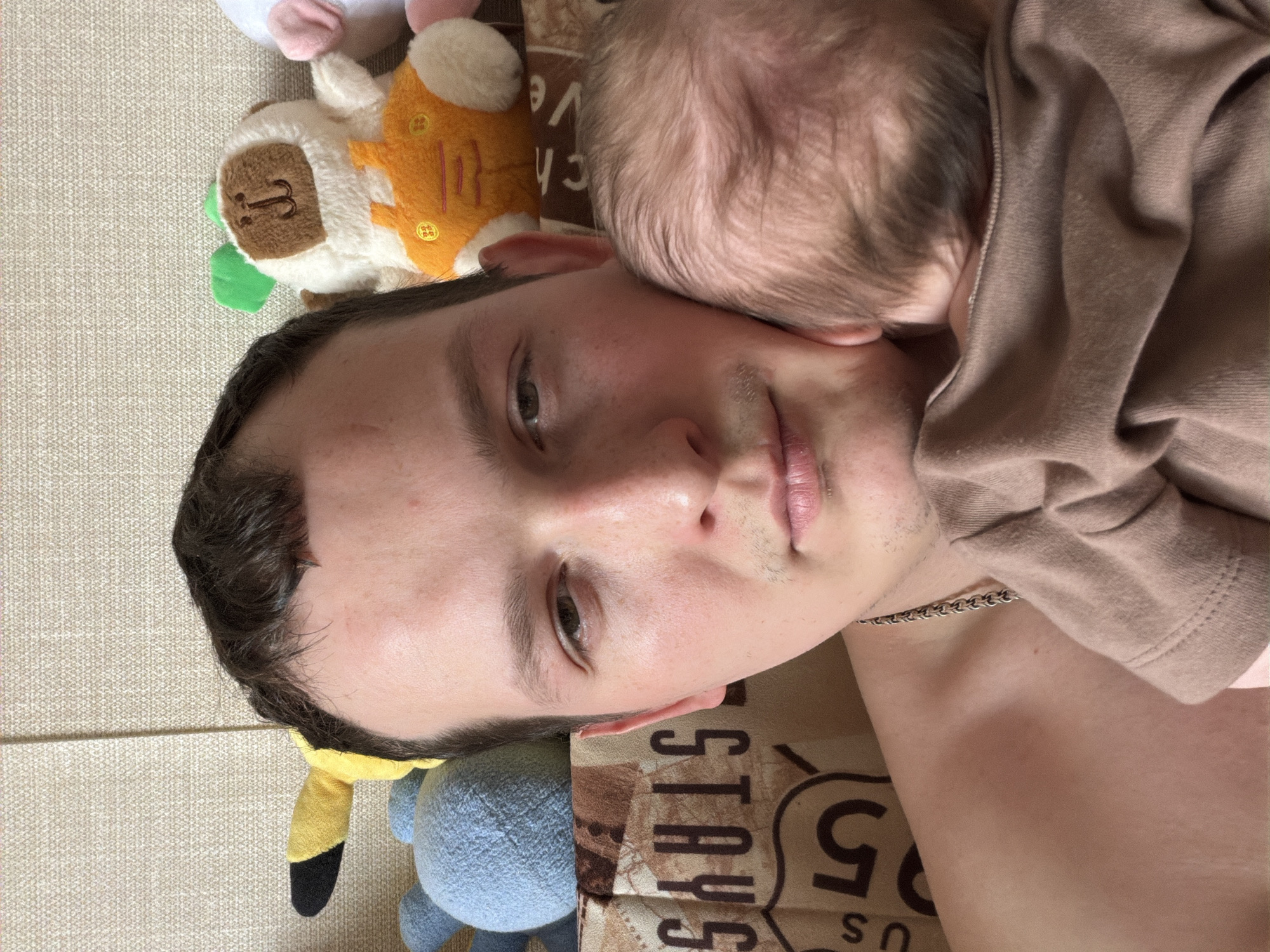 An adult male holds an infant indoors. The male, appearing to be in his late 20s or early 30s with short dark hair, light skin, and stubble, looks directly at the camera. He wears a metallic chain around his neck and is shirtless or wearing a low-cut garment. An infant, with light brown hair and wearing a light brown top, is held against the man's left shoulder, its head resting against his cheek and facing away from the camera. The background features several stuffed animals: a yellow and black plush, a blue spherical plush, and a white and brown monkey in orange overalls. Behind the man's left shoulder, a patterned fabric displays partial text reading "STAYS" and "US" next to a large "5." The immediate background is a light beige textured fabric, consistent with an indoor domestic setting. The image itself provides no verifiable visual information to confirm a specific location within Chaikovsky, Russia.