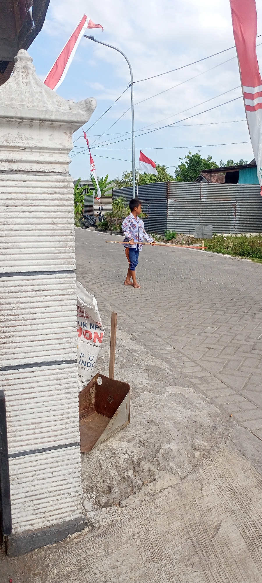 The image depicts a street scene in Gempolklutuk, Indonesia, with a young boy standing on a paved road holding a long, thin object. He is wearing a patterned shirt and blue shorts. In the background, a corrugated metal fence lines the street, and houses are visible beyond it. Indonesian flags, red and white, are prominently displayed on poles along the street. To the left of the frame, a textured white pillar and a white bag are in the foreground. A metal dustpan and a wooden stick are also visible on the ground near the bag. A motorcycle is parked on the side of the road behind the boy. The sky is partly cloudy.