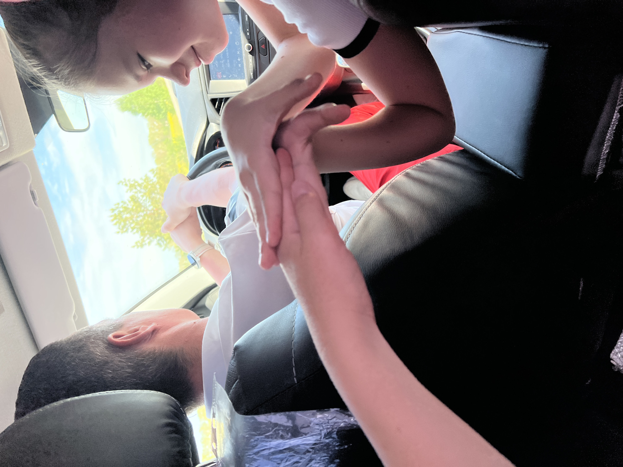 Inside a vehicle, a woman with fair skin is positioned above a man in the driver's seat. The woman is looking down at the man and is holding his hands. The car's dashboard is visible in the background. The environment outside the vehicle includes trees and a blue sky. The location is unspecified.