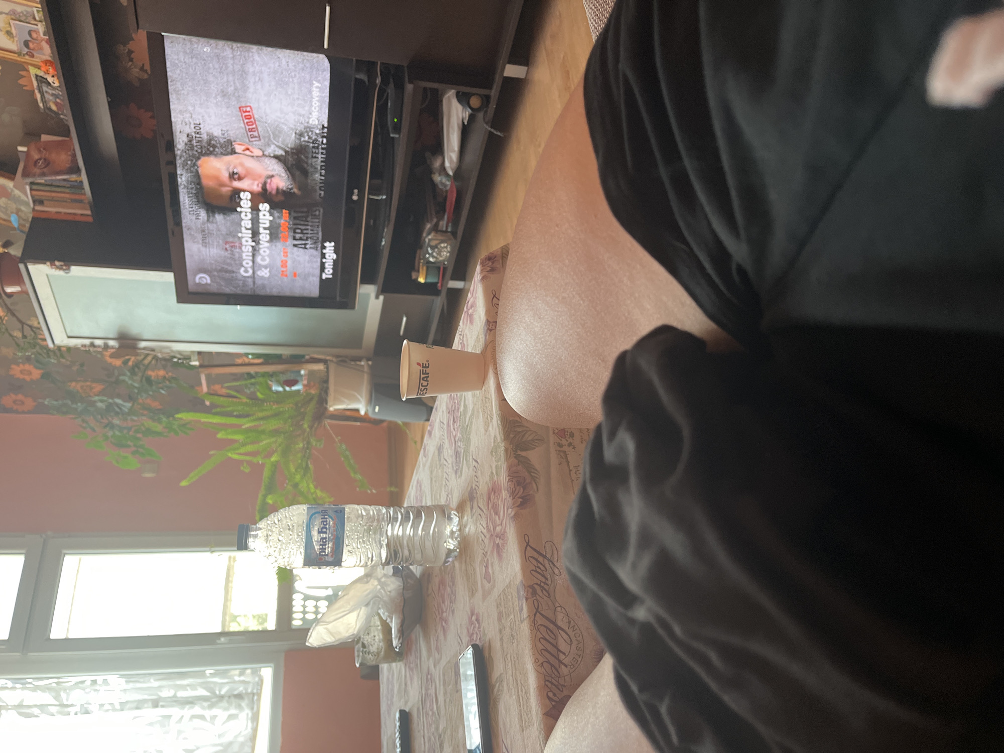 This image captures a first-person perspective from an individual reclining in a residential living room, consistent with an interior setting in the city of Plovdiv, Bulgaria. The foreground reveals the person's upper thighs and lap, covered by dark fabric shorts.
A rectangular table, draped with a light-colored tablecloth featuring a floral and textual pattern (including "Love Letters"), is positioned directly in front of the viewer. On the table, from left to right, are a dark-colored smartphone, a TV remote control, a small glass or bowl containing a yellowish substance and wrapped in white plastic, a transparent plastic bottle of "Горна баня" (Gorna Banya) mineral water, and a light-colored paper cup with "NESCAFÉ" branding.
In the midground, a dark-colored flat-screen LG television is centered on a matching dark TV stand. The television screen displays a program banner titled "Conspiracies & Coverups" from the Discovery channel, featuring a bearded man and indicating "21.00 CET Tonight." Below the TV, the stand's open shelves hold various items, including books and a device with a visible green indicator light. Above the TV, a shelf contains additional books and framed pictures.
To the left of the television, a tall, dark cabinet with a light-colored door is visible. To the right of the TV, on the floor, a potted green plant is situated. Another potted plant is partially visible further left in the room. The room features a wall covered with floral wallpaper (orange flowers on a lighter background) behind the TV and to its right. A window with sheer white curtains, allowing ample daylight into the room, is on the left side, above a light orange-peach wall section. The floor visible beneath the TV stand is a light-colored wood or laminate. No other individuals are explicitly visible in the scene.