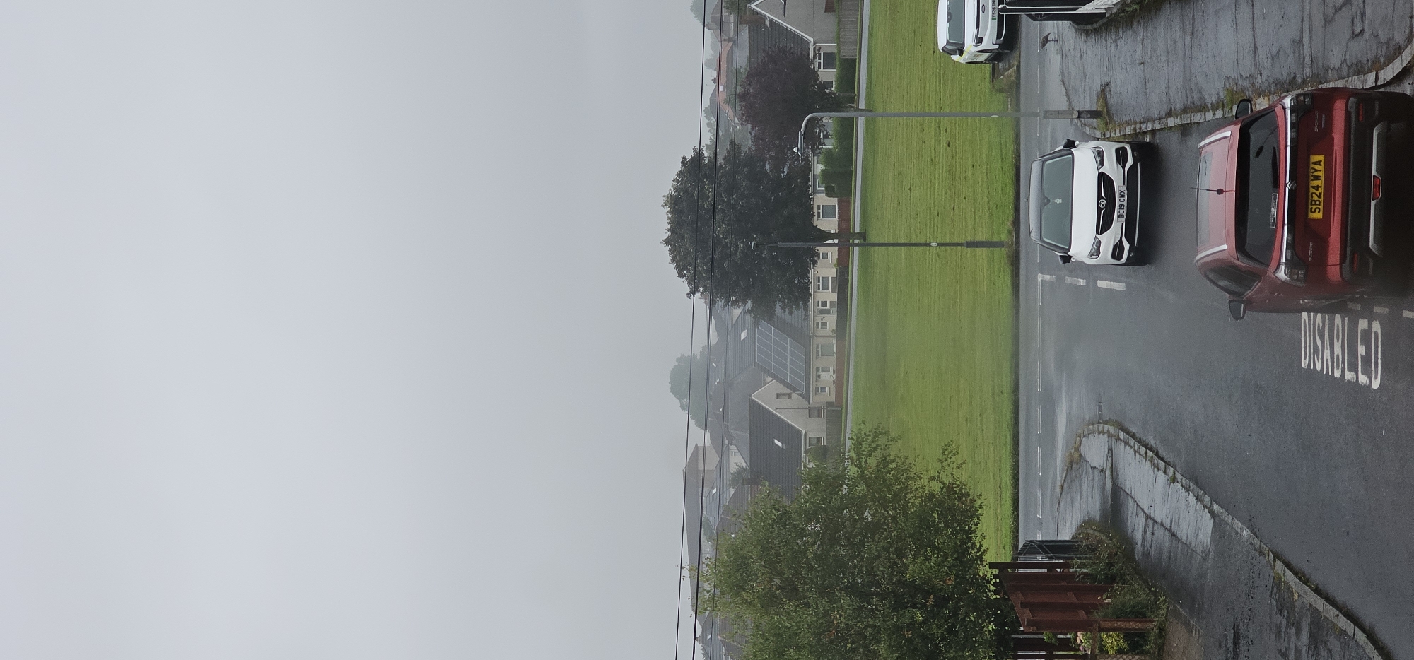 The image depicts a view of a residential street in Kilwinning, United Kingdom, on an overcast day, with visible rain creating wet surfaces. In the foreground, a red Suzuki Vitara SUV is parked on the street, with the word "DISABLED" stenciled in white on the wet asphalt behind it. Further up the street, a white Vauxhall Grandland X is parked. Beyond the street, a large, manicured green space is visible, bordered by houses and trees. Some houses in the background are equipped with solar panels. Power lines stretch across the scene, and a lamppost stands near the green space. A bird is visible on the pavement near the white car. The sky is heavily overcast, obscuring the horizon and lending a muted quality to the scene.