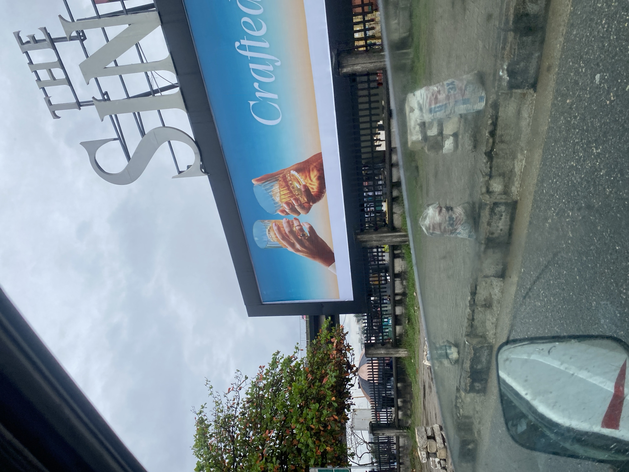 This image captures a scene on Victoria Island, Nigeria, featuring a large billboard advertising a beverage. The billboard prominently displays two hands holding glasses filled with amber liquid, suggesting a whiskey or similar spirit. The text "Crafted" is visible on the billboard, alongside the brand name which appears to be "THE SIN". The billboard is positioned above a dark railing, with a street and a sidewalk in the foreground. On the sidewalk, several concrete blocks and bags of what appear to be cement or sand are stacked. A large tree with green and reddish leaves is situated to the left of the billboard. The sky is overcast, indicating a cloudy day. The perspective of the photograph suggests it was taken from inside a vehicle, with the side mirror visible in the lower left corner.