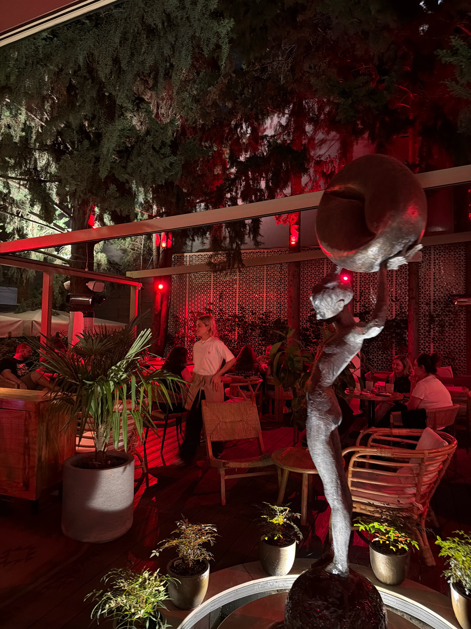 The image captures an outdoor dining area at night, illuminated by red ambient lighting. A bronze sculpture of a figure holding a sphere above its head stands in the foreground. Surrounding the sculpture are potted plants and chairs made of wood and wicker. In the background, several patrons are seated at tables, engaged in dining and conversation. One person, likely a server, is standing and looking towards the right. The setting appears to be a restaurant or bar in Tbilisi, Georgia, with lush trees overhead and a patterned screen or wall in the background. The overall atmosphere is dimly lit and intimate, with the red lights casting a warm glow on the scene.