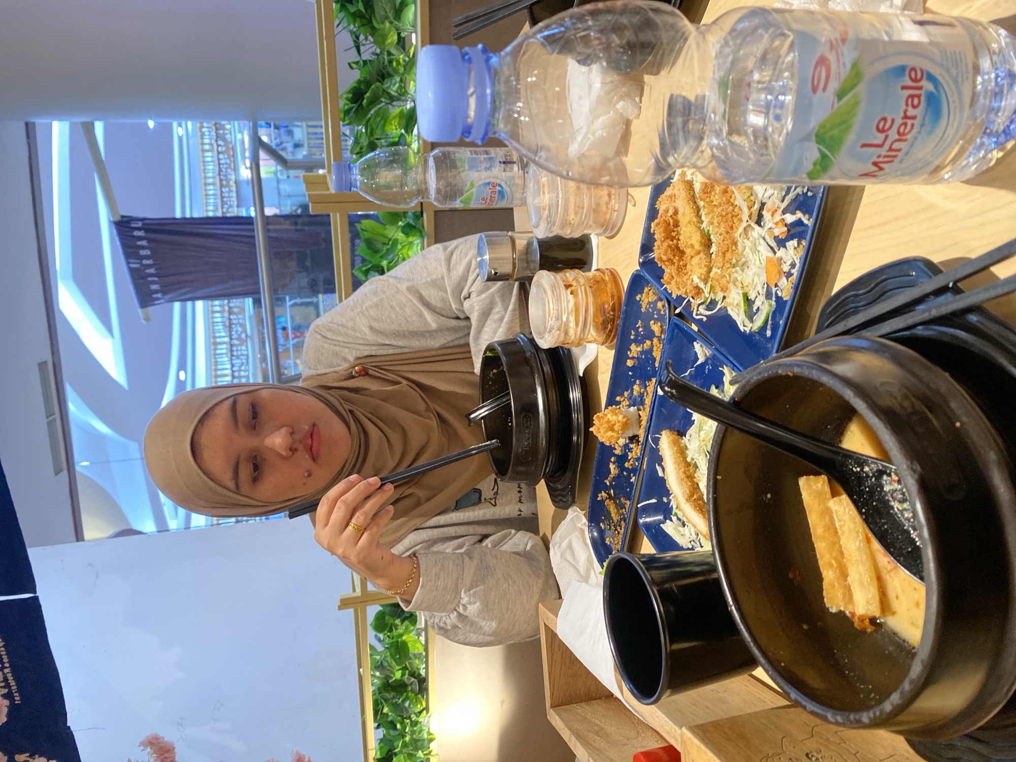 The image depicts a young woman wearing a hijab and a gray sweatshirt sitting at a table in what appears to be a restaurant in Banjarbaru, Indonesia. She is looking down at her food, holding a pair of chopsticks. On the table in front of her are several blue plates with food, including what looks like fried chicken or fish, and some shredded vegetables. There are also bowls, bottles of water, and small containers of condiments. In the background, there is a banner with Indonesian text that reads "BANJARBARU". The restaurant appears to be indoors, with natural light coming from above and a view of the city beyond. The overall scene suggests a casual dining experience.