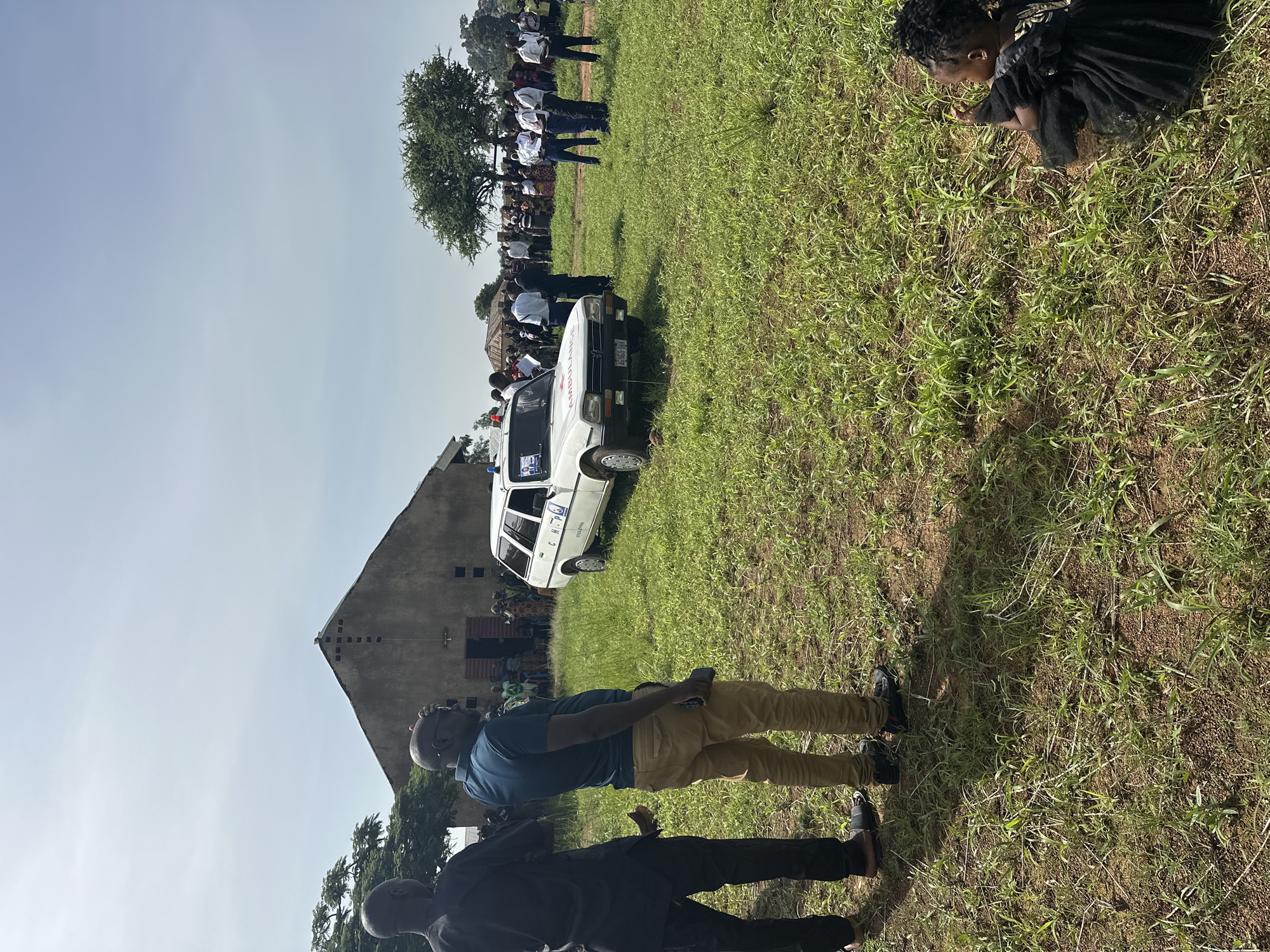 The image depicts an outdoor gathering under bright daylight, likely in a rural or semi-urban setting in Panyam, Nigeria. The scene is dominated by a wide, grassy field with sparse green growth, suggesting dry ground. The sky is clear and blue, indicating sunny weather.
In the foreground, two men stand facing away from the camera. One wears a blue polo shirt and tan trousers, holding a dark object (possibly a phone) in his hand. The other, partially obscured, is in dark clothing. Their shadows are cast long on the ground, suggesting mid-morning or late afternoon.
Further back, a white sedan-style vehicle, possibly a Peugeot, is parked in the grass. It is clearly marked as an "AMBULANCE" on its front hood and also has a red cross symbol visible. On its side, the text "CHPO" and a license plate reading "S 586 MV" can be seen.
Behind the ambulance, a crowd of people is gathered, some standing in rows, many wearing white shirts, suggesting a uniform or specific attire for an event. They are standing near a large tree with green foliage. In the far background, a rustic building with a distinctive sloped roof and small square openings is visible, potentially a community center or a place of worship.
To the right, separate from the main crowd, a person, possibly a child, dressed in a dark, ruffled outfit, is crouching in the grass, looking down. The overall impression is of a community event or public assembly, possibly involving health services given the presence of the ambulance.