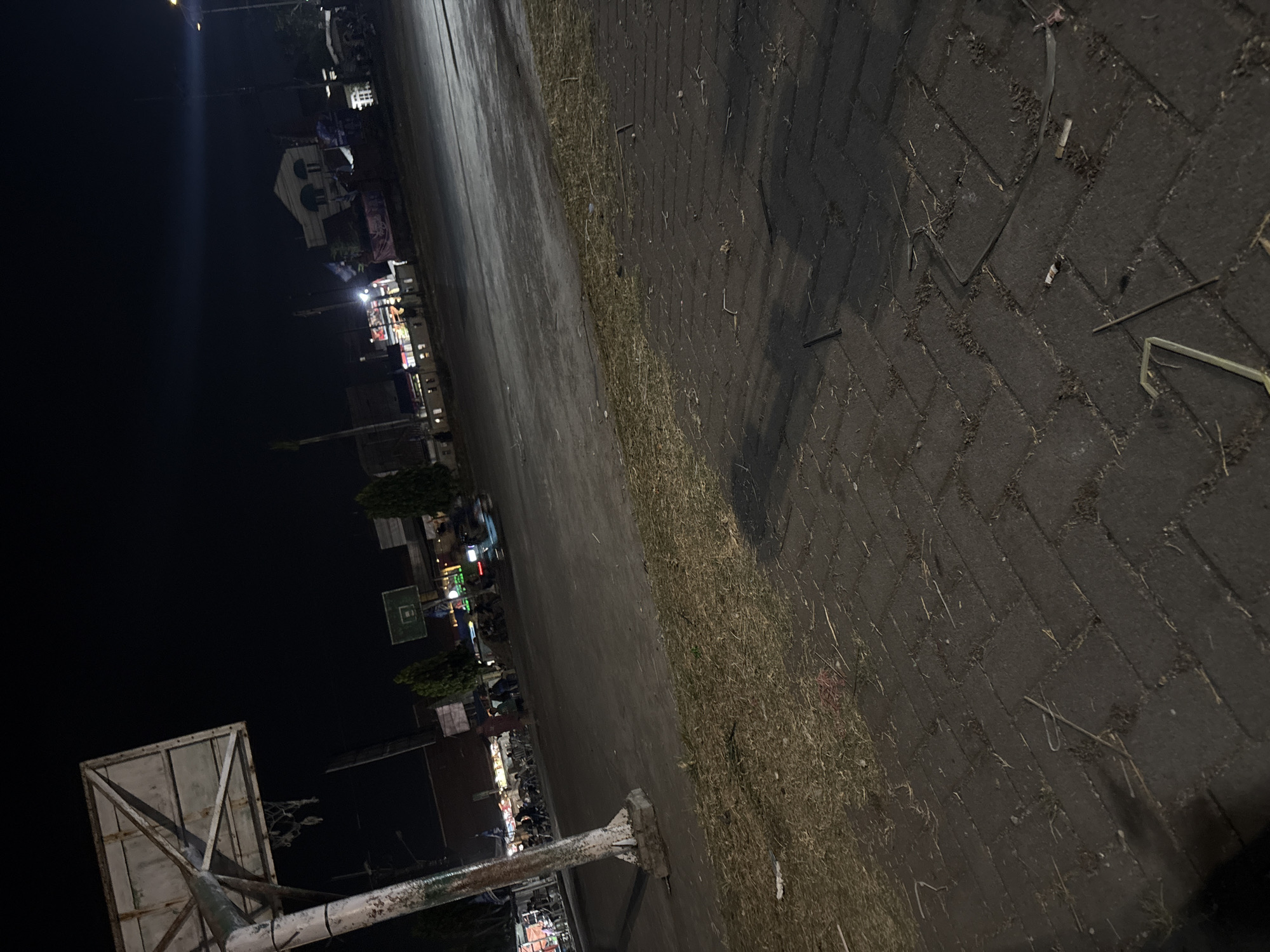 This is a low-light, wide-angle photograph taken at night, likely in the city of Palimanan, Indonesia. The foreground features a patterned brick sidewalk with scattered dry grass and debris. To the left, a weathered basketball hoop structure stands prominently, its pole showing signs of rust and wear. Beyond the sidewalk is a paved area that appears to be a street or open space, with a grassy verge separating it from the sidewalk. In the mid-ground, several trees are visible, and behind them, buildings with illuminated windows and signs suggest a commercial or residential area. Several motorcycles are parked or in motion, indicating activity. Further in the background, a building with a distinctive white facade and arched windows is illuminated, and flags are visible. The overall impression is of a public space at night, possibly a recreational area or a roadside marketplace, in Palimanan.