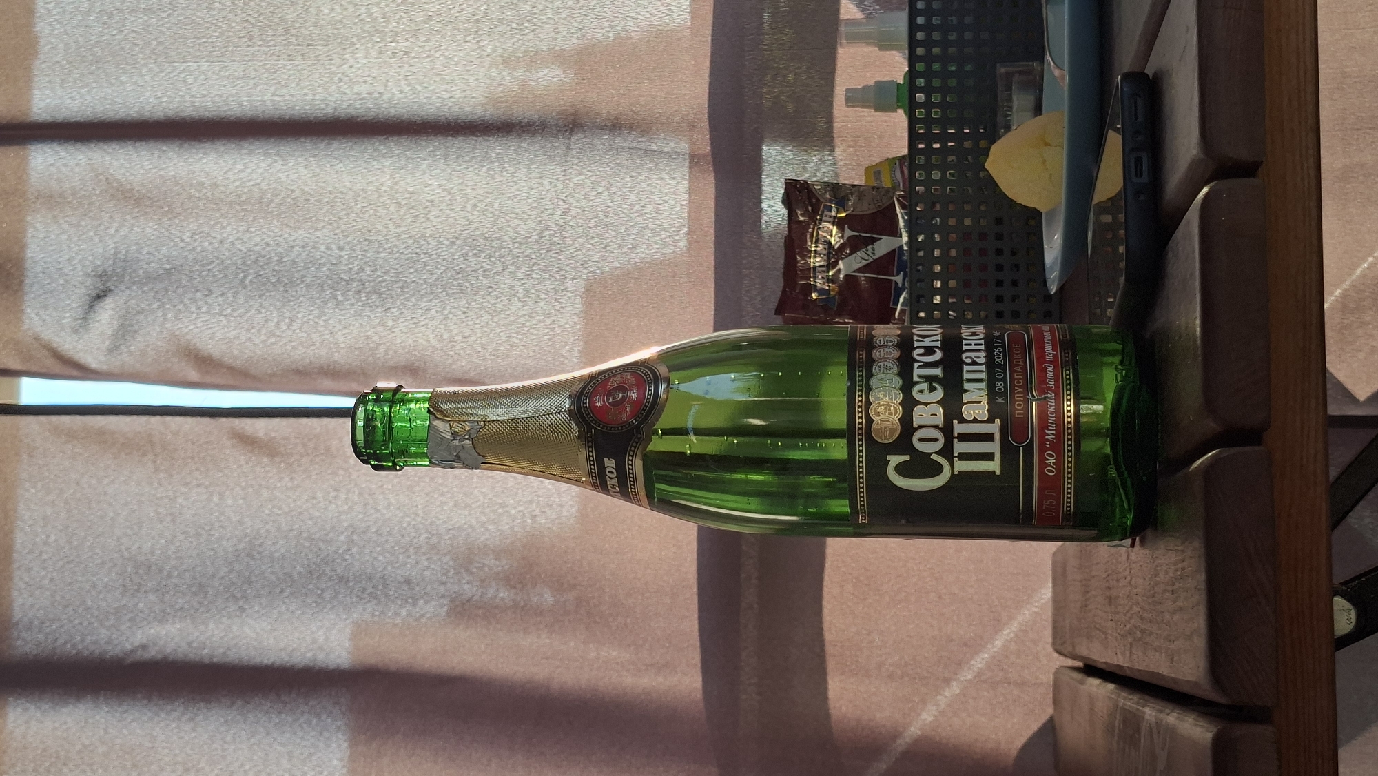 A green glass bottle of "Soviet Champagne" stands on a wooden table. The bottle has a gold foil cap and a label that reads "Советское Шампанское" (Soviet Champagne). Below this, it says "ПОЛУСЛАДКОЕ" (semi-sweet), the volume "0.75 л" (0.75 L), and the manufacturer "ОАО "Минский завод игристых вин"" (JSC "Minsk Sparkling Wine Plant"). A date "К 08.07.2026 17:46" is also visible.
In the background, sheer pink curtains are drawn, with a bright light source behind them. To the right of the bottle, on a shelf, is a foil packet of snacks, two small spray bottles, and a plate with a piece of yellow food, possibly cheese or butter. A smartphone lies on the table in front of the plate.
The setting appears to be an outdoor picnic or dining area, possibly in a dacha or cafe. The presence of the specific manufacturer on the bottle confirms the location as Belarus, specifically linking to Minsk, but the wider context suggests a picnic setting outside of Minsk city proper. Given the presence of Minsk Sparkling Wine Plant, the image could have been taken anywhere in Belarus where this product is sold and consumed. However, without further visual cues, specifying a precise location within Myadzyel, Belarus is not possible based on the image alone.