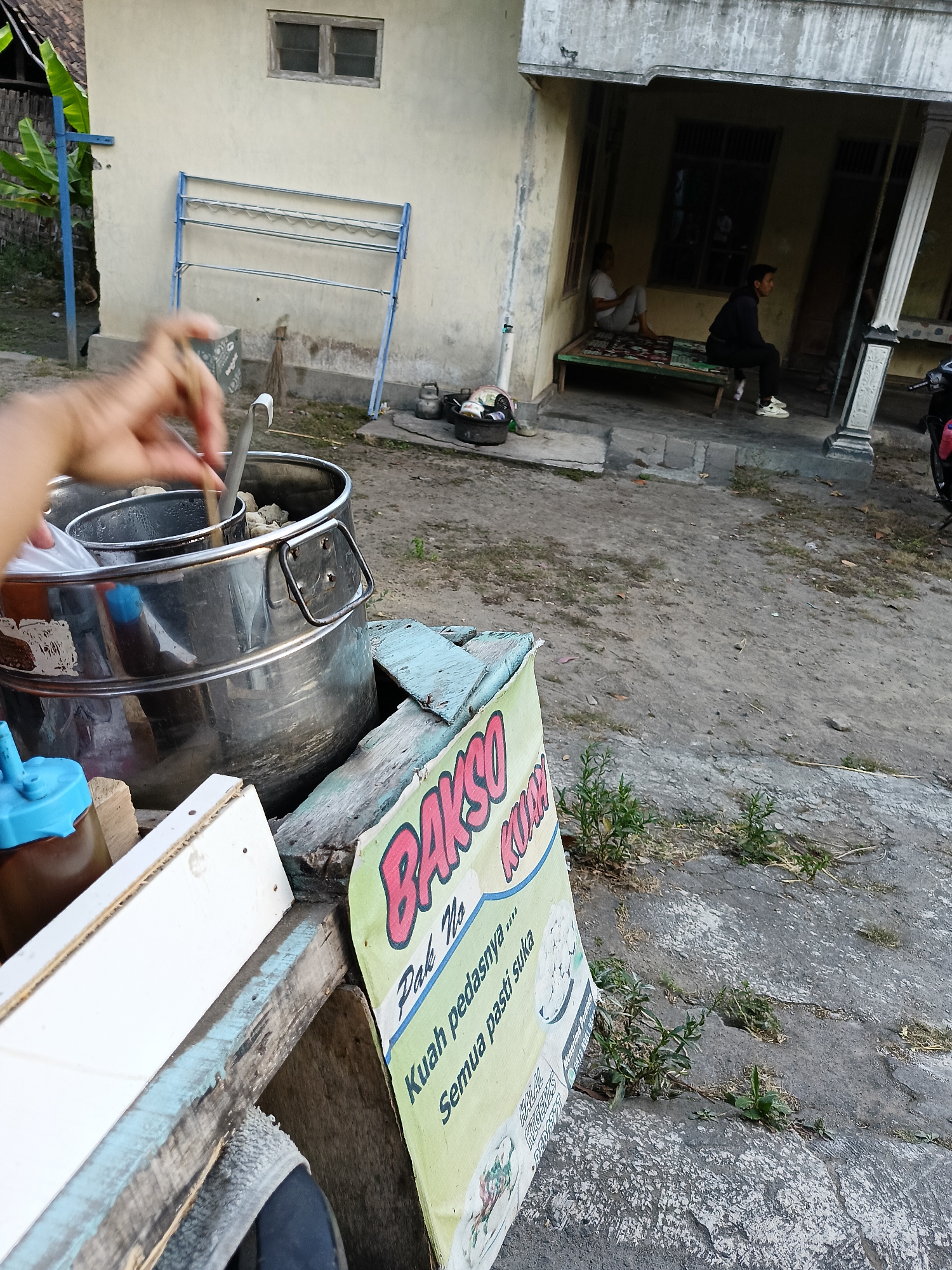 In Borangan, Indonesia, a close-up shot captures a food stall with a large stainless steel pot. A hand is seen stirring the contents of the pot with a utensil. To the right of the pot, a sign with text "BAKSO KUAH" and "Pak No" is visible, with smaller text below reading "Kuah pedasnya..." and "Semua pasti suka". In the background, a simple house with an open-air veranda is visible. Two men are seated on a raised platform under the veranda, with one appearing to be an adult and the other a younger individual. A motorbike is parked to the right of the veranda. The ground is a mixture of dirt and cracked pavement.