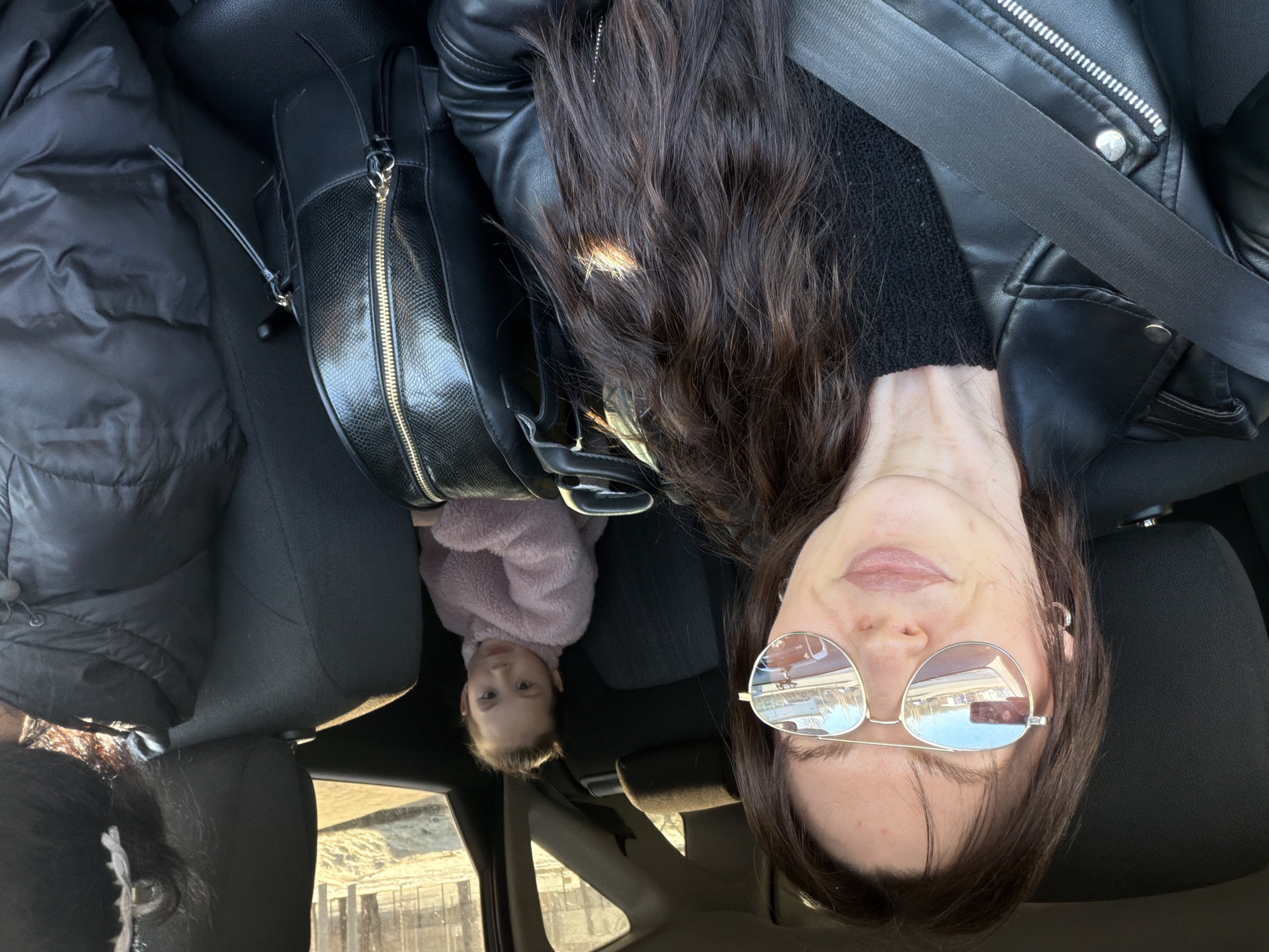 Captured from inside a car, the image shows an adult woman and a baby in the backseat. The woman, wearing a black leather jacket and sunglasses, appears to be positioned with her head near the car's roof. Her long, dark hair cascades around her. A black backpack sits behind her. The baby, dressed in pink, is seated in the middle of the backseat. A winter coat is also visible on the left. Through the car window, a low structure with a fence is visible, suggesting a location within Perm, Russia. The time of day appears to be daytime.