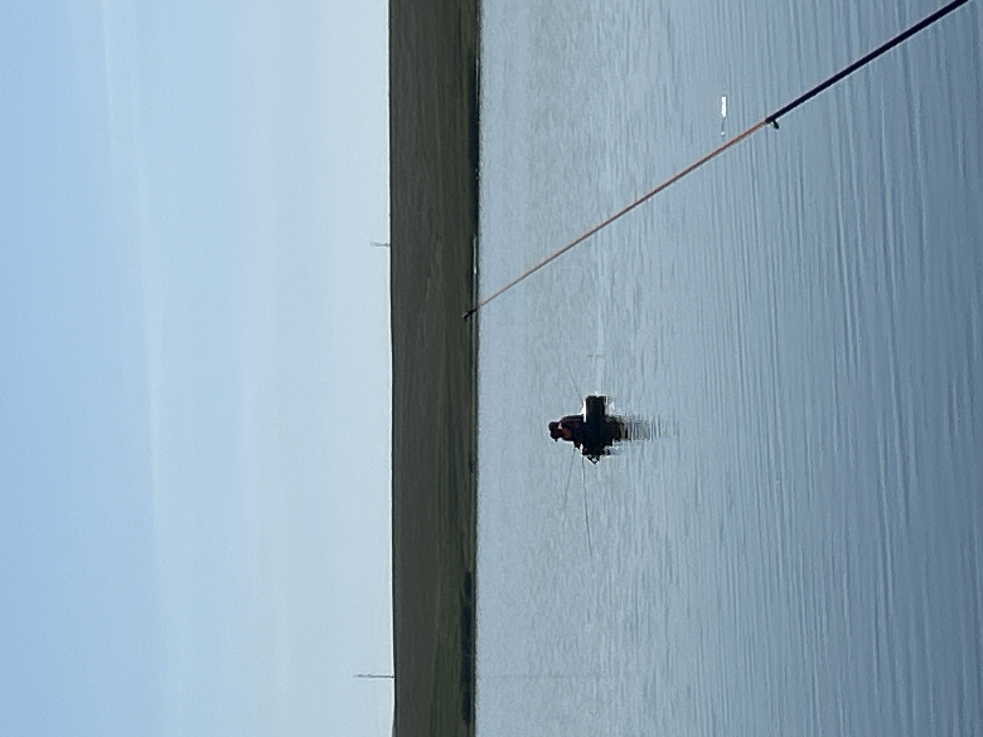 The image depicts a serene, calm body of water, likely a lake or reservoir, under a clear, light blue sky. In the middle distance, a small, dark-colored raft or floating platform is visible. On the raft, two figures are seated, facing away from the viewer. One figure appears to be wearing a life jacket. They are fishing, as evidenced by fishing rods extending from the raft. The raft is casting a slight reflection on the water's surface. In the background, a long, green, grassy embankment or dam stretches horizontally across the frame, with a faint, tall, slender structure visible on its crest in the far distance, possibly a utility pole or marker. The foreground of the image is dominated by a fishing rod, held by an unseen person, its orange and black line extending diagonally across the water. The water itself is calm, with subtle horizontal ripples distorting the reflections of the sky and distant features. The scene suggests a quiet, recreational activity taking place in a natural, open environment. The specific location in Matraevo, Russia, is not visually identifiable from the image itself, but the overall setting is consistent with a rural or semi-rural area featuring a body of water.