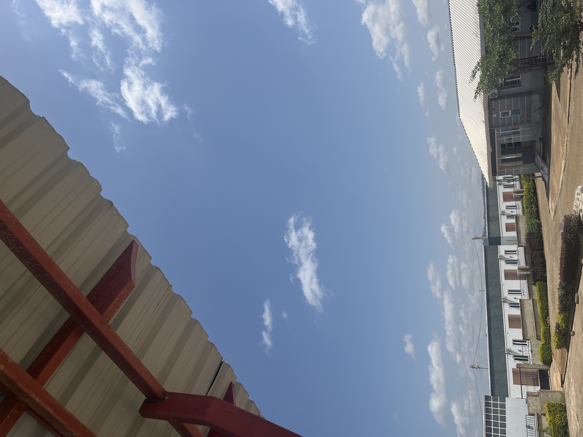 The image captures a view of a building complex and a corrugated metal roof under a bright, clear blue sky with scattered white clouds. The building on the right is a multi-story structure with a white roof, several windows, and air conditioning units visible. Some sections of the building are grey, and there is a tree with green foliage in the foreground. The corrugated metal roof on the left is supported by red metal beams. The scene appears to be outdoors in a sunny environment, suggesting it is daytime. There is no visible text in the image. The overall impression is one of a typical day in a populated area, possibly residential or commercial, with a focus on the architecture and the sky. The orientation of the image is portrait.