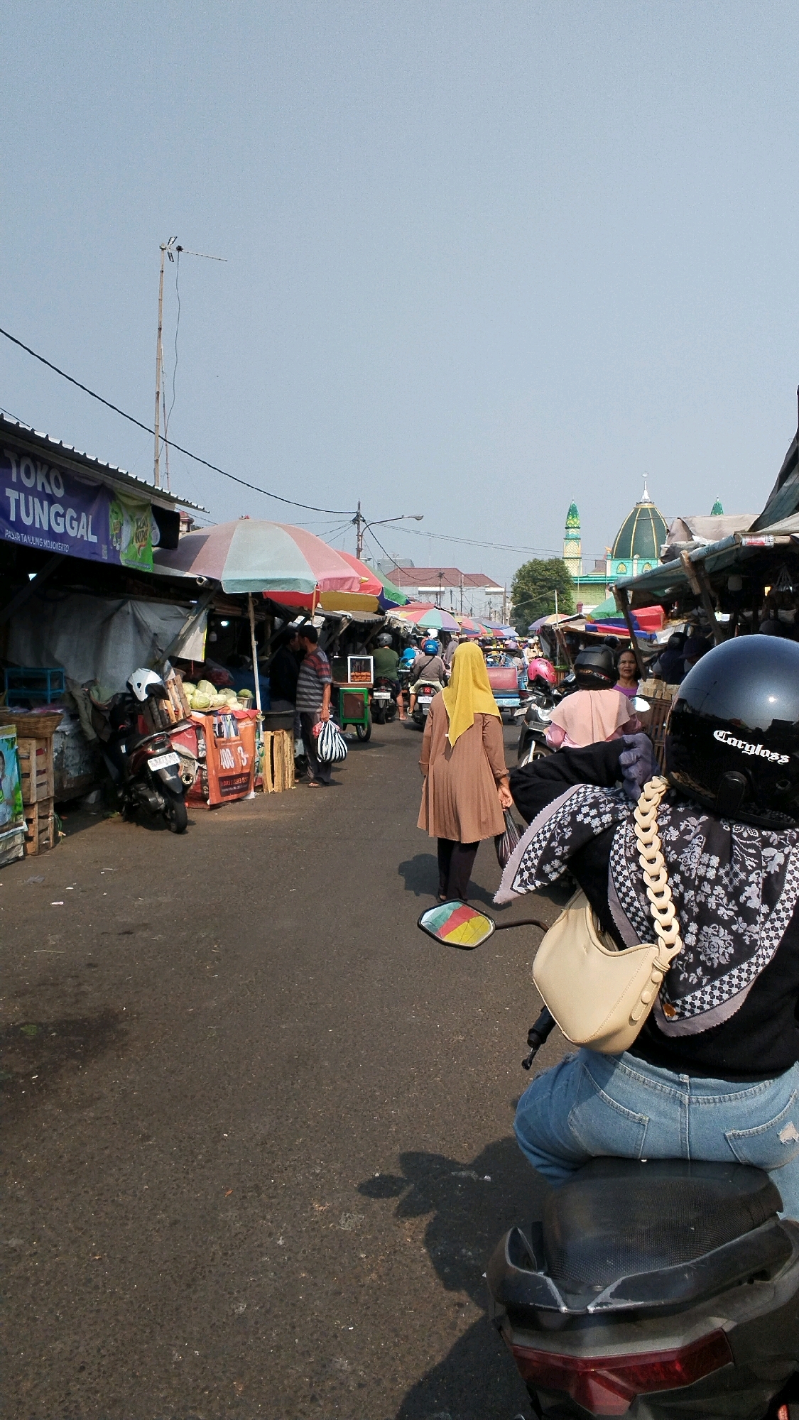 The image depicts a busy street market scene in Mojokerto, Indonesia. The foreground shows the back of a person on a motorcycle, wearing a black helmet with "Carloss" printed on it, a black and white patterned scarf, and blue jeans. The motorcycle is parked on an asphalt road. In the middle ground, a woman in a brown headscarf and long dress walks away from the camera, carrying a white bag. Several stalls line the street, covered by colorful umbrellas, selling various goods, including produce. Motorcycles and people are seen moving through the market. In the background, a mosque with a green dome and minarets is visible. The sky is clear and bright, suggesting it is daytime. The overall atmosphere is one of a bustling local marketplace.