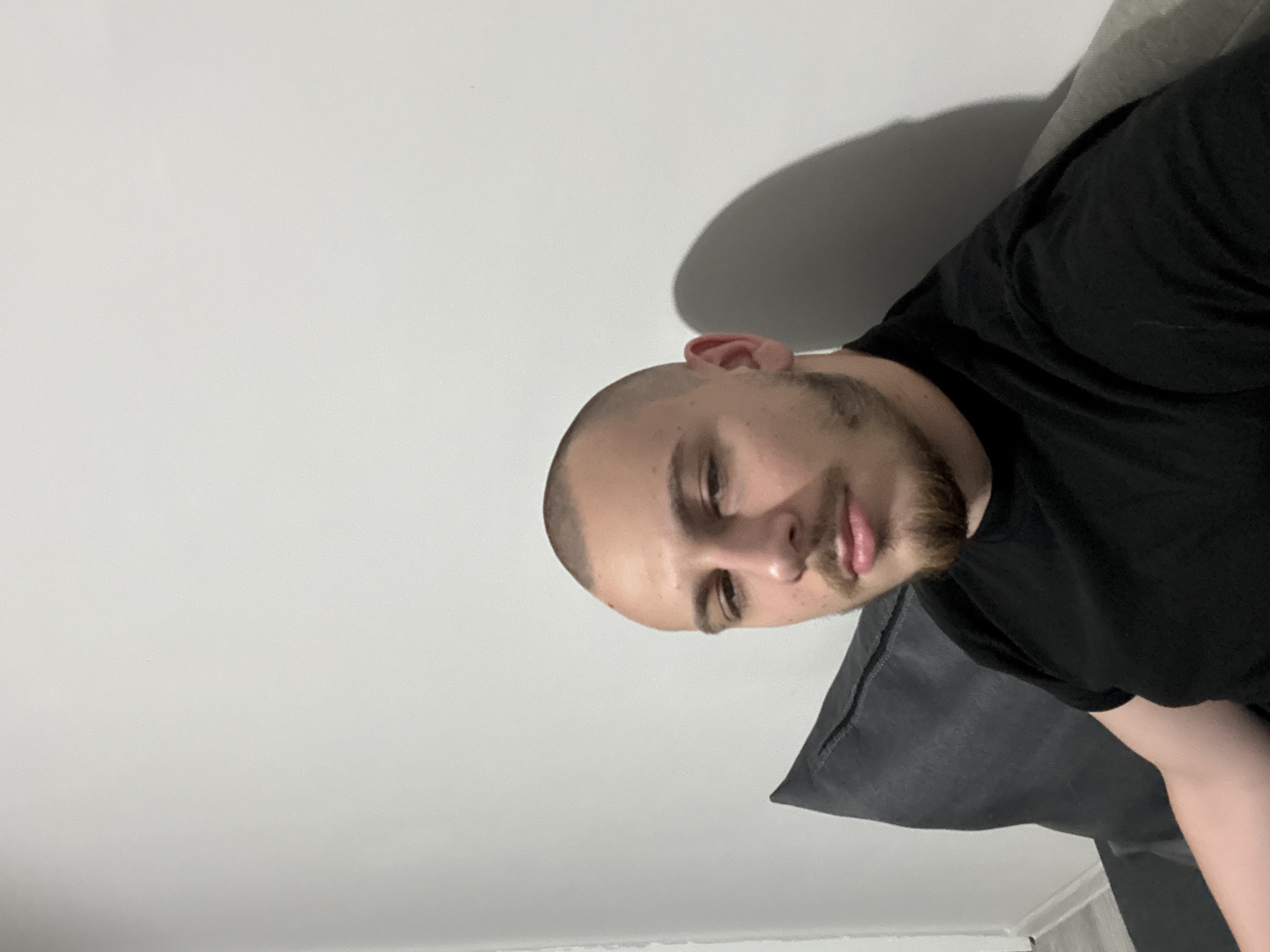 A man with a shaved head and a short beard is lying on a dark gray couch or cushion. He is wearing a black shirt. The background is a plain, light gray or white wall. There is a shadow on the wall above his head. The location is not identifiable as Stara Zagora, Bulgaria based on the visual information provided.