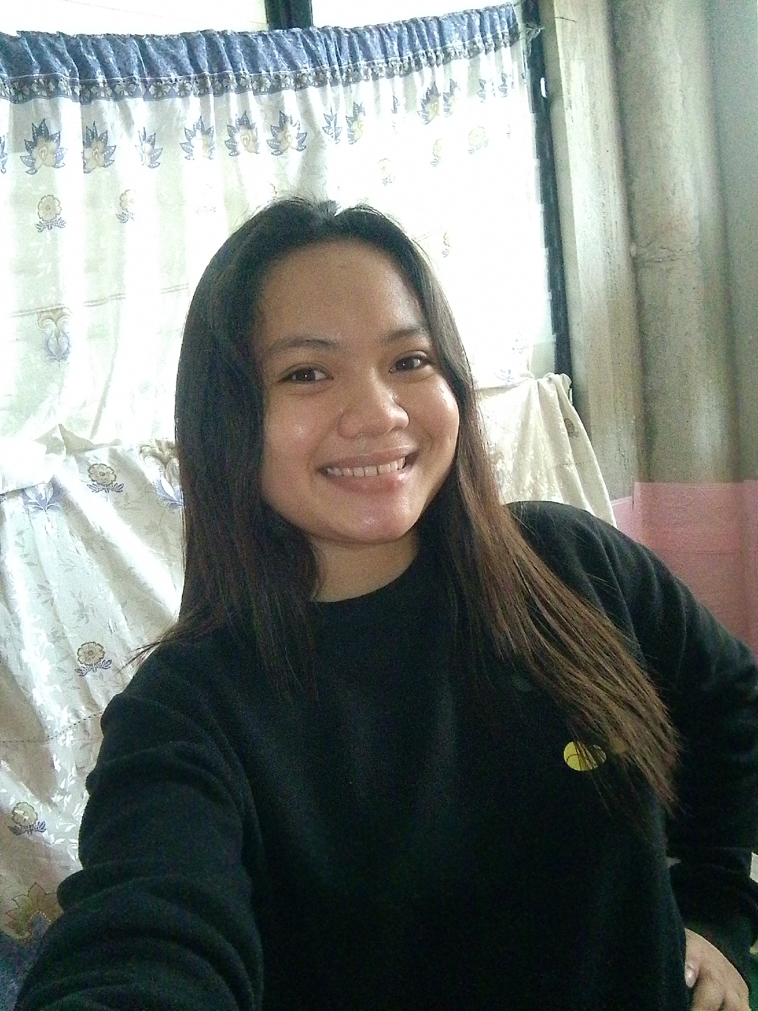 A medium shot taken from a slightly high angle shows a young woman with long, dark brown hair smiling and looking directly at the camera. She is wearing a black crew neck sweatshirt. A small, yellow circular emblem is visible on the left side of her chest. Behind her, a patterned curtain with floral motifs in blue and light yellow can be seen. To the right of the curtain, a textured wall with horizontal lines is visible. The lighting in the scene appears to be natural daylight.
