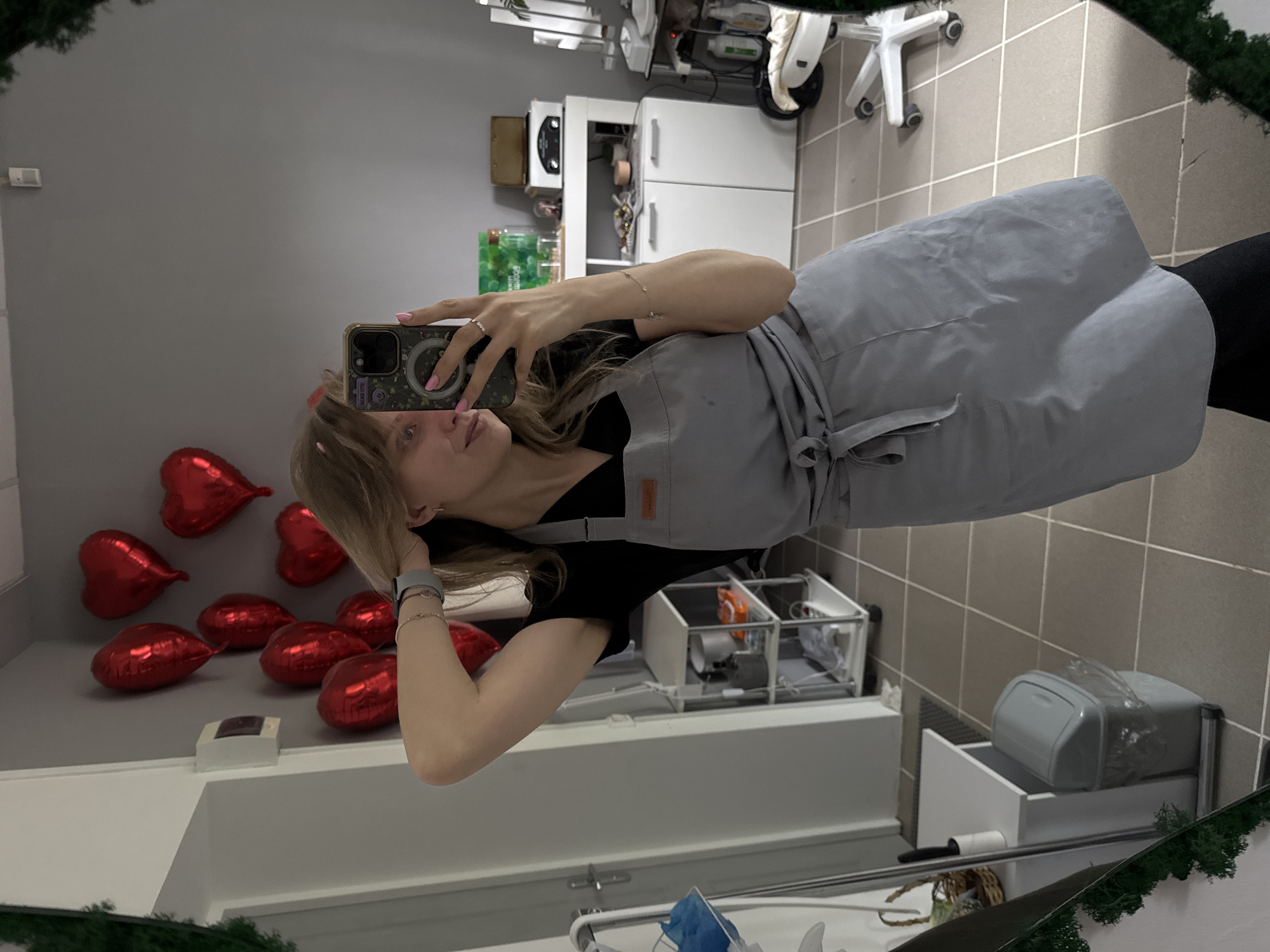The image shows a young woman taking a selfie in a mirror. She has blonde hair and is wearing a black t-shirt and a grey apron. She is holding a smartphone with a decorative case in her right hand. Behind her, there is a wall with several red heart-shaped balloons attached to it. There is also a white cabinet, a white trolley, and other salon equipment visible in the background. The floor is tiled. Given the presence of salon equipment and decorations, the location appears to be a beauty salon in Izhevsk, Russia.