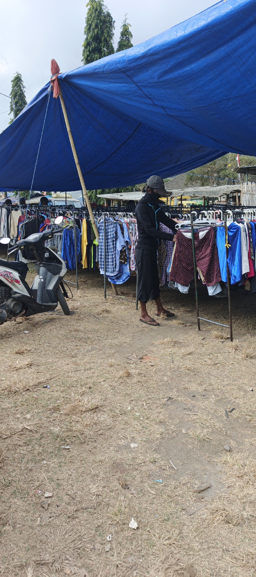 A person stands under a blue tarp at an outdoor clothing market, likely in Puger, Indonesia. The person, wearing a black long-sleeved shirt, black shorts, a grey cap, and a face mask, is examining a rack of clothing. A white scooter is parked to the left of the frame. The ground is covered in dry grass and dirt. Racks of various colored shirts and pants are displayed. In the background, there are trees and some buildings. The sky is overcast.