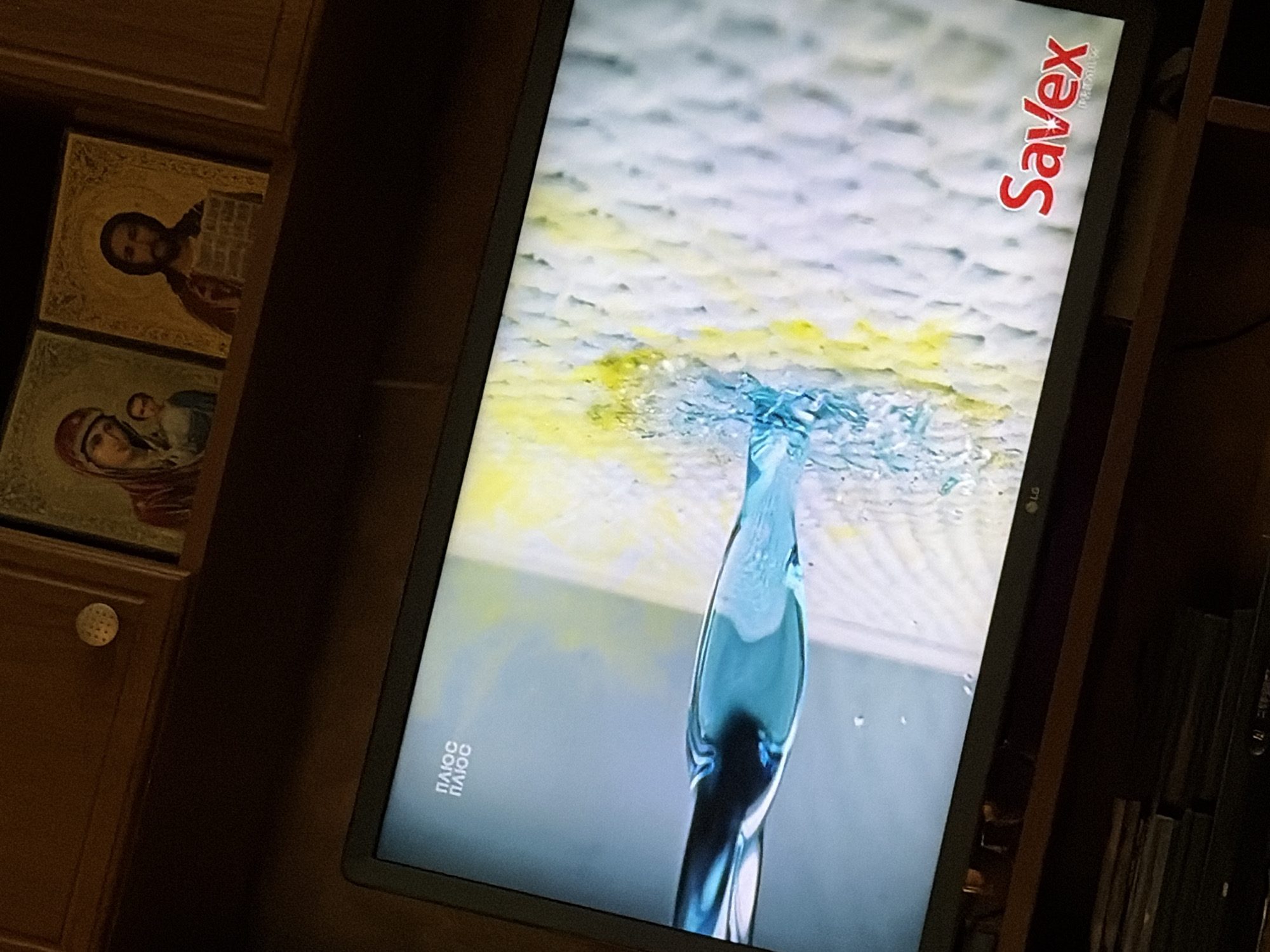 A television screen displays a commercial with blue liquid being poured onto a textured white surface with yellow streaks. In the upper left corner of the screen, the word "ПЛЮС" is displayed twice in white text. In the lower right corner of the screen, the word "Savex" is prominently displayed in red text with a smaller white logo underneath. The television is placed on a wooden shelf, with stacks of what appear to be CDs or DVDs below it. Above the television, on a wooden cabinet, are two framed religious icons. The icon on the left depicts a woman and child, and the icon on the right shows a man. The overall scene suggests an indoor setting, likely a living room or entertainment area. The image is taken from a slightly elevated angle, looking down at the television.