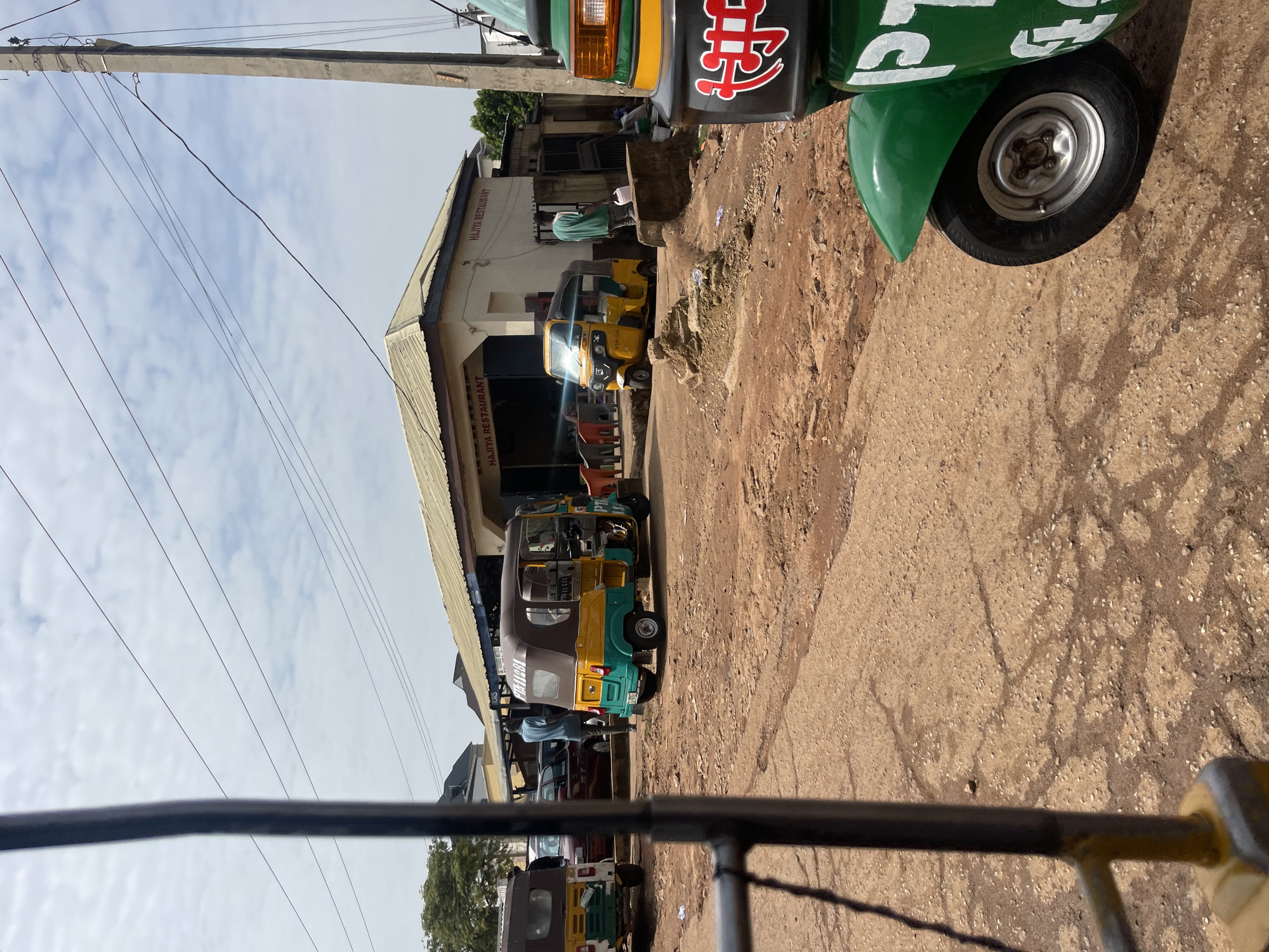 In the city of Jos, Nigeria, a busy street scene unfolds in front of "Hajiya Restaurant." The restaurant, a single-story building with a light-colored exterior and a corrugated metal roof, displays its name prominently on both sides of the facade. Several auto-rickshaws, also known as Keke Napeps, are parked or driving in the dusty, unpaved street. One auto-rickshaw, a yellow and green model, is parked directly in front of the restaurant, its passenger compartment visible. Another green and yellow auto-rickshaw is parked to the left, with its rear facing the camera. A third, primarily yellow auto-rickshaw, is seen from the side. A man in a blue shirt and dark trousers walks away from the restaurant towards the parked auto-rickshaws. Further down the street, a person in a green shirt and dark pants is seen near the entrance of what appears to be another establishment. The environment is characterized by dry, reddish-brown earth and sparse vegetation. Overhead, power lines crisscross the sky, supported by a concrete utility pole. The sky is partly cloudy, with a bright, overcast day.