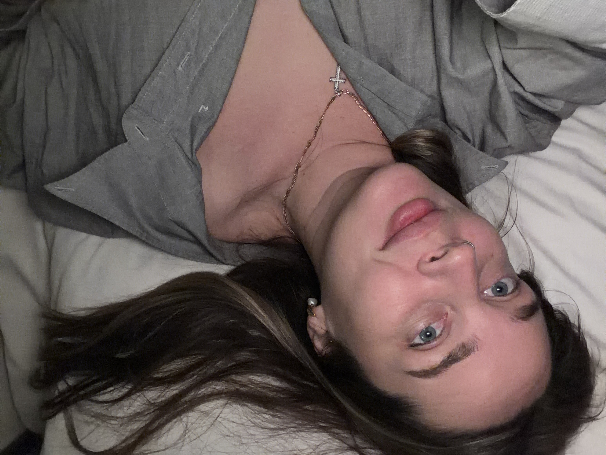 A young woman is depicted lying on her back, looking upwards towards the camera, suggesting a selfie or a close-up portrait. Her head is slightly tilted, and her long, dark brown hair is spread out on a white or light-colored surface, likely a bed. She wears a loose-fitting, light grey collared shirt, which is unbuttoned near the neckline.
Adorning her neck is a delicate gold-toned chain necklace featuring a silver-colored inverted cross pendant. She has a small pearl earring in her visible left ear and a subtle silver hoop nose ring in her right nostril. Her blue eyes are open and gaze directly at the viewer, with well-defined dark eyebrows. Her lips are full and slightly parted, carrying a soft, natural hue and a hint of a smile, contributing to a relaxed or pensive expression.
The setting appears to be indoors, likely a bedroom, with the white bedding serving as a simple background. The lighting is soft and even, without harsh shadows, hinting at an indoor, possibly evening or low-light environment. There are no explicit location cues within the image itself to confirm the provided context of Moscow, Russia. No visible text is present in the media.