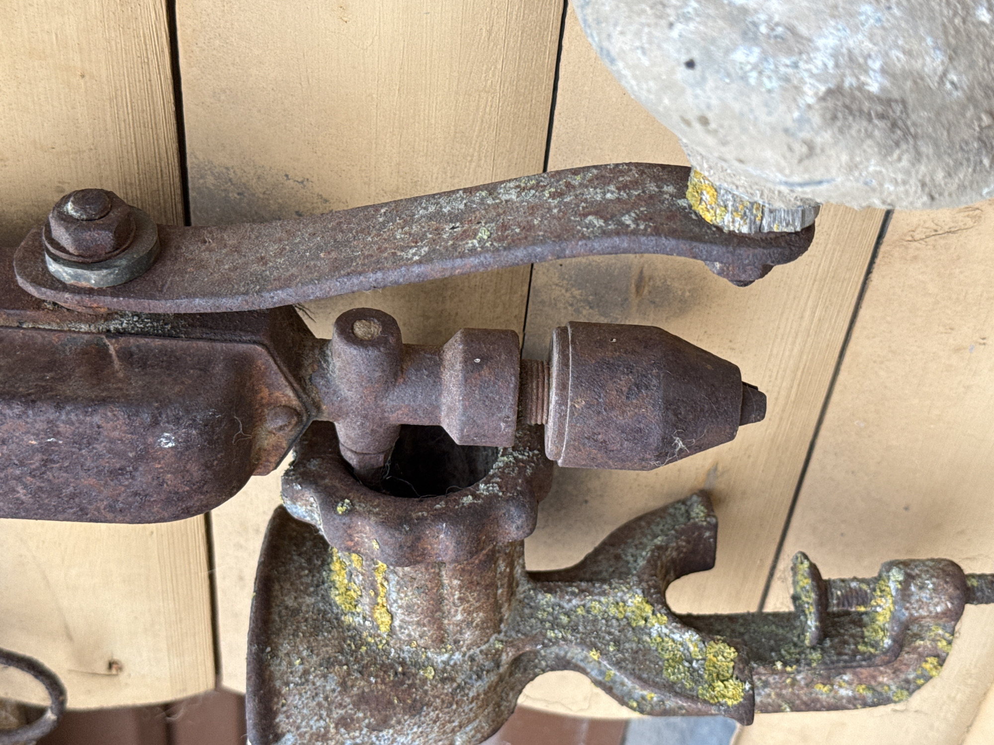 A close-up, eye-level shot captures a rusted, antique meat grinder clamped to a light-colored wooden wall. The grinder, a mix of dark brown rust and hints of green lichen, is positioned in the foreground, its various components, including the hopper, the clamp, and the crank mechanism, are clearly visible. To the right of the grinder, a large, rough-textured, light gray object, possibly a stone or concrete block, partially obscures the lower right corner of the frame. The wooden wall, with its horizontal planks and visible grain, serves as the backdrop, suggesting an outdoor setting. The lighting is natural and appears to be from the side, casting subtle shadows and highlighting the textures of the aged metal and wood. The scene is devoid of any human presence. This scene is located in Vikraičiai, Lithuania.