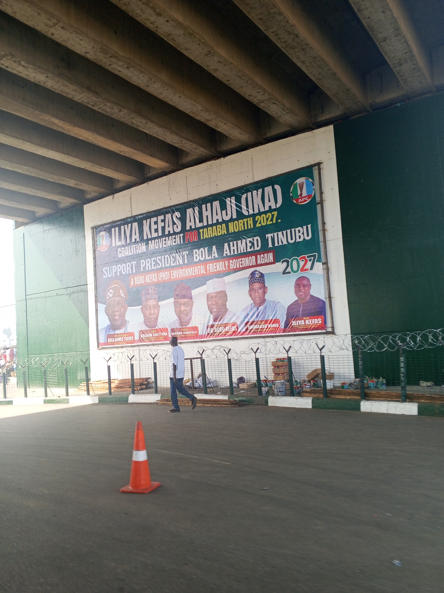 The image shows a banner displayed on a wall under an elevated concrete structure, likely a flyover or bridge, in Jalingo, Nigeria. The banner features portraits of five men, with text indicating support for "ILiya Kefas Alhaji (Ika) Coalition Movement for Taraba North 2027." It also mentions "SUPPORT PRESIDENT BOLA AHMED TINUBU & AGWU KEFAS (PHD) ENVIRONMENTAL FRIENDLY GOVERNOR AGAIN 2027." The APC logo is visible on the banner.
In the foreground, a man in a light blue shirt and dark trousers walks away from the camera on an asphalt surface. An orange traffic cone with a white reflective band is positioned in the middle ground. Behind the traffic cone and the man, a wire fence with barbed wire runs along the base of the wall. Various cans of paint, likely for the banner or the wall, are visible behind the fence, along with wooden planks. The environment appears to be an outdoor public space, possibly a roadside or a market area in Jalingo. The elevated concrete structure creates a shaded area over the scene.