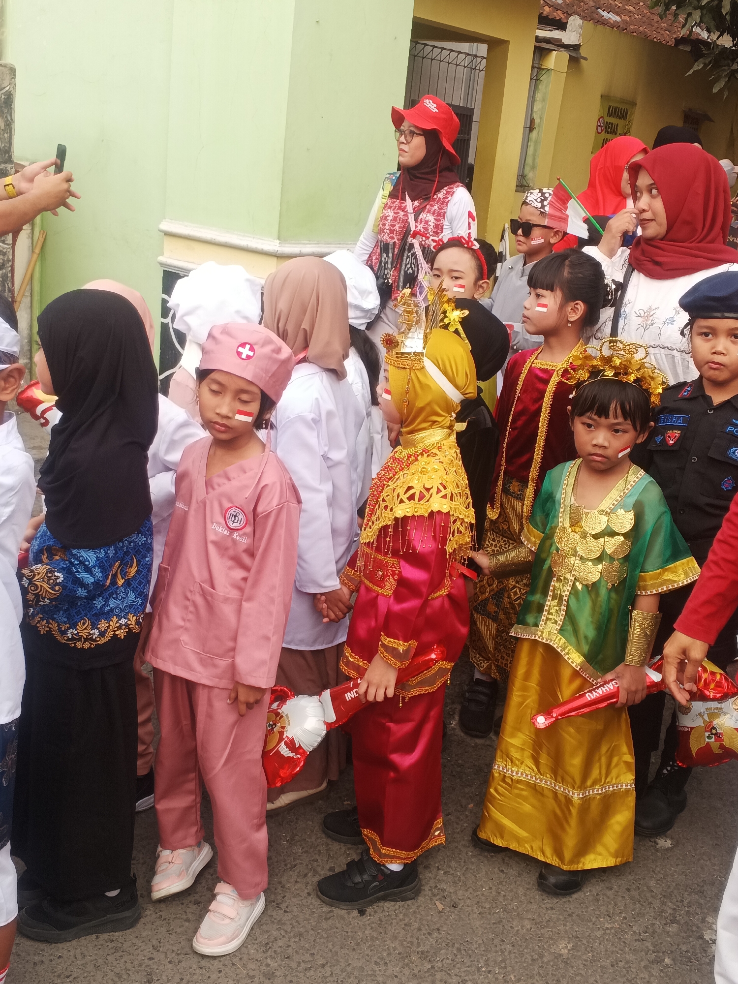 A group of children and adults, some in traditional Indonesian attire and others in costumes, are gathered on a paved street in Sokanegara, Indonesia. The children are dressed in various outfits, including what appears to be a doctor's uniform, a traditional golden costume, and outfits with Indonesian flag colors. Several children have Indonesian flag markings on their faces. Some adults are also wearing red and white, suggesting a patriotic event. A person on the far left is holding a smartphone, seemingly taking a picture or recording. The background shows buildings with a light green and yellow facade, indicating an urban or semi-urban setting. The overall atmosphere appears festive, possibly related to a national holiday or celebration.