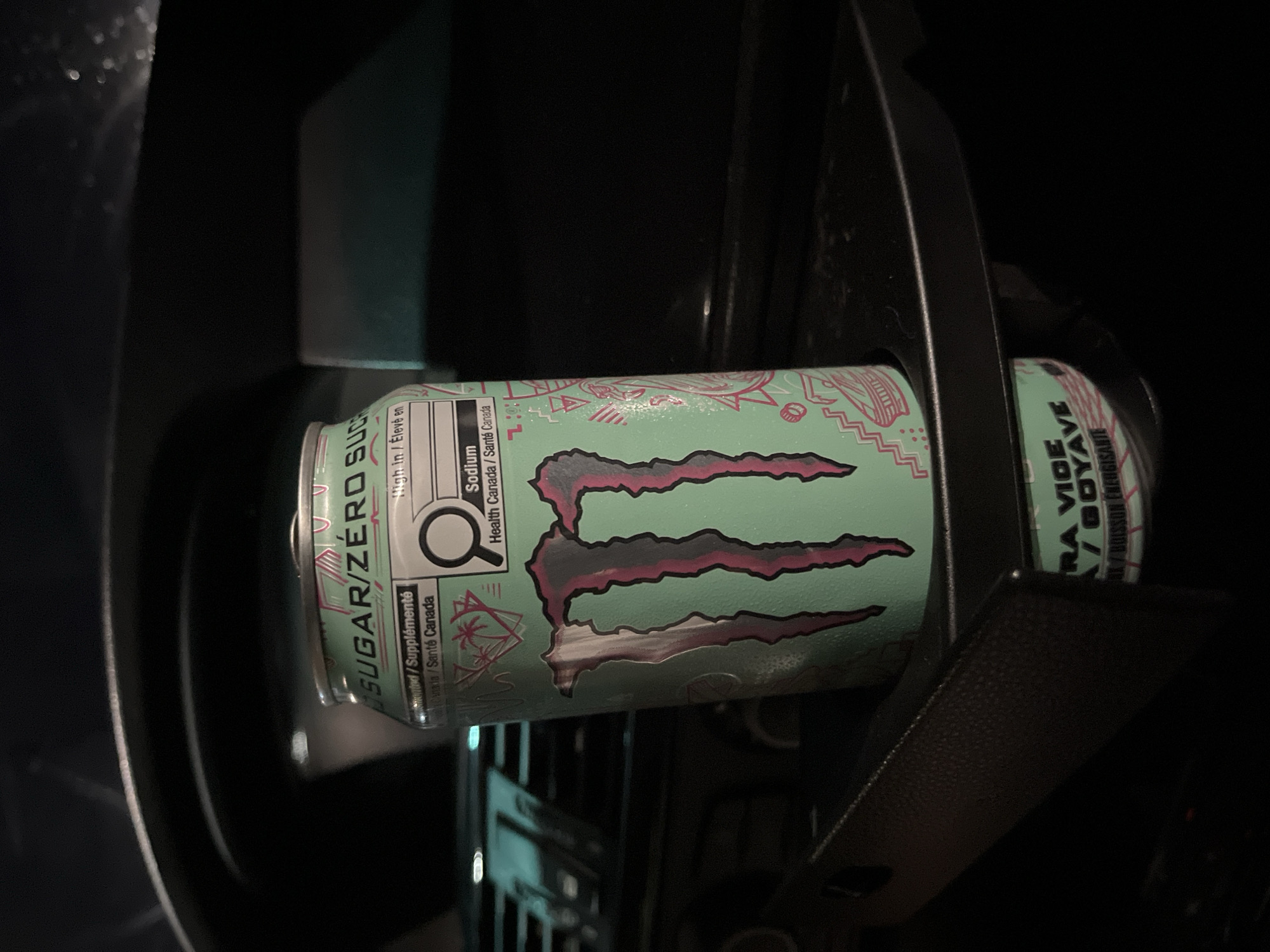 A can of Monster Energy Ultra Paradise sugar-free drink is nestled in a car's cup holder. The can is light blue-green with the Monster claw logo outlined in pink and black. The can's label includes nutritional information in both English and French, noting "SUGAR/ZERO SUCRE" and "Sodium" along with "Health Canada/Santé Canada" and a warning label "High in/Élevé en". Part of the label reads "RA VICE / BOYAVE", with the full flavor likely "GUAVA VICE". The cup holder is surrounded by dark car interior components. There's no indication of the city of Shumen, Bulgaria.