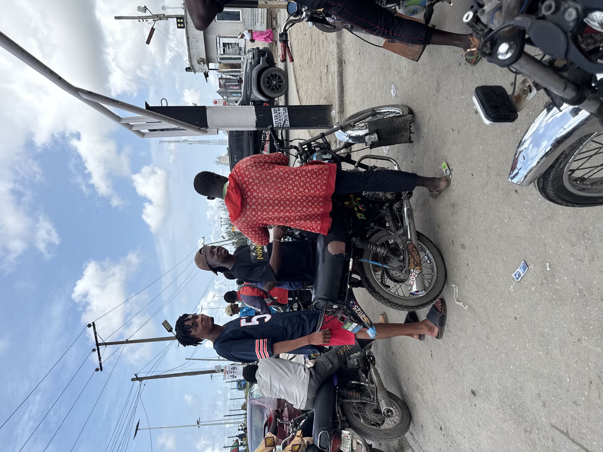 This media file captures a vibrant daytime street scene in an urban or semi-urban setting, likely in Lagos, Nigeria, given the context. The sky is bright blue with scattered white clouds, indicating clear weather.
The primary subjects are several men, with three prominently featured in the foreground and midground, interacting with or standing near motorcycles. One man, wearing a distinct red jacket with a repeating pattern, is positioned on a motorcycle. Beside him, another man in a black t-shirt and a backward baseball cap stands near a second motorcycle. Further to the left, a third man with dreadlocks, dressed in a blue and white jersey with the number "5" and red shorts, stands next to a third motorcycle, wearing sandals. Other people are visible further down the street in the background, including one person wearing pink.
The scene appears to depict a bustling roadside or possibly a checkpoint. Motorcycles are a significant element, parked or idling on the paved surface. In the background, a grey car or SUV is visible, and various buildings line the street. Utility poles with extensive wiring crisscross the frame, and a white barrier arm, possibly for a gate or a road closure, extends into the upper left of the image.
Visible text includes multiple signs and markings. On a black and white striped pole, there's a clear sign stating "SALES GIRL/BOY NEEDED" with a contact number "09134567649", along with fragmented text like "MURYAN DA" and "TACT". In the background, a partial sign reads "PLEASE ALL THOSE GO", "COMPLEX MUST W", "DESTRIAN &", and "KINDLY C", suggesting instructions for a specific area. The years "2025" and "2026" are visible on parts of one motorcycle, possibly indicating registration or model year. The general atmosphere is one of routine daily activity.