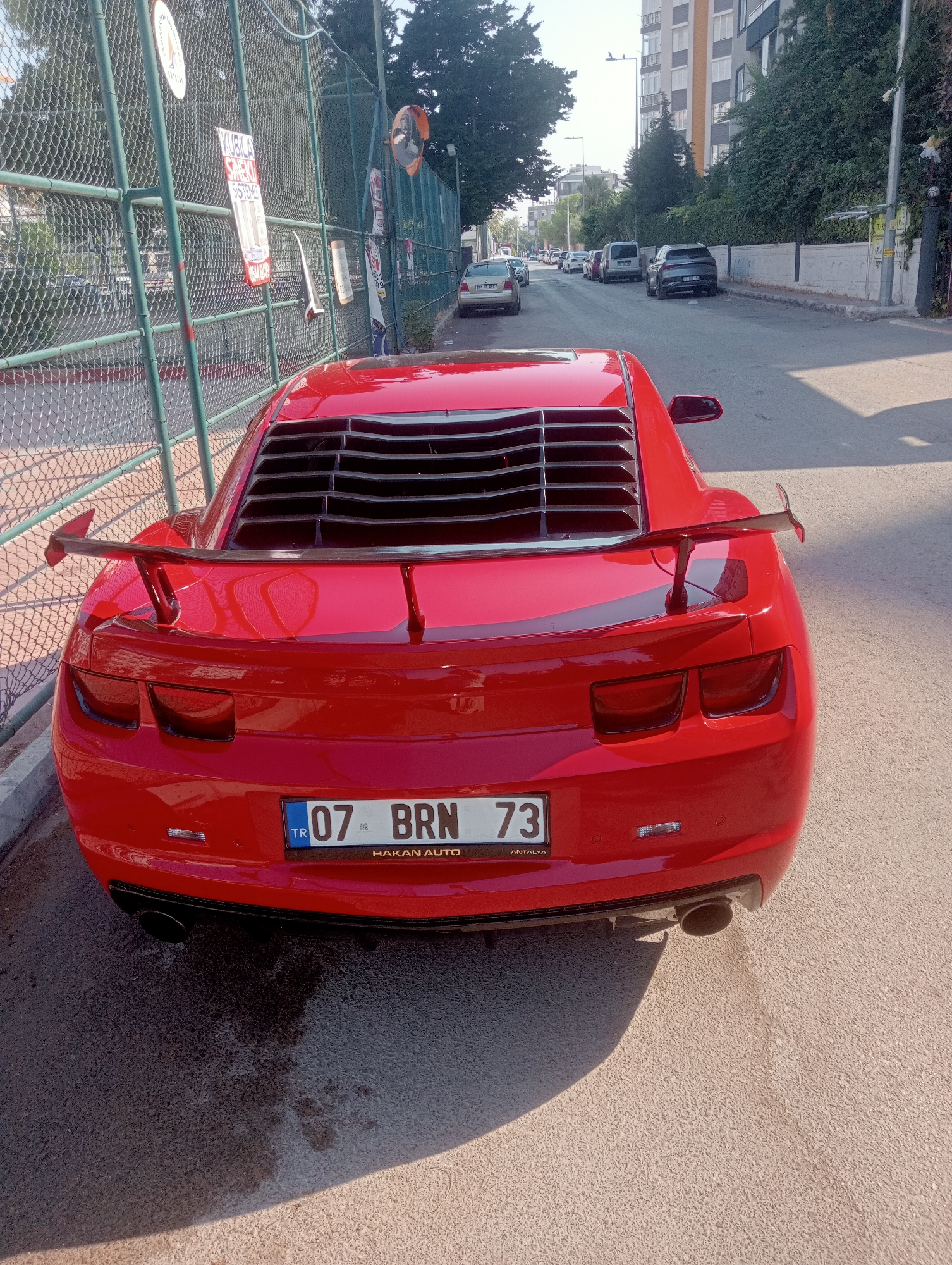 In a sun-drenched street scene in Antalya, Turkey, a bright red Chevrolet Camaro is parked adjacent to a chain-link fence. The car, viewed from the rear, features a prominent spoiler and louvers covering the rear window. Its license plate reads "07 BRN 73," with "ANTALYA" printed below. To the left, the fence is adorned with various posters and notices. Beyond the fence, a red curb borders a sidewalk, and further back, a row of parked cars lines the street, with a multi-story building visible in the distance. The street surface appears to be asphalt, with some wet patches and tire marks visible on the ground in front of the car. The lighting suggests it is daytime, with shadows cast by the fence and the car.