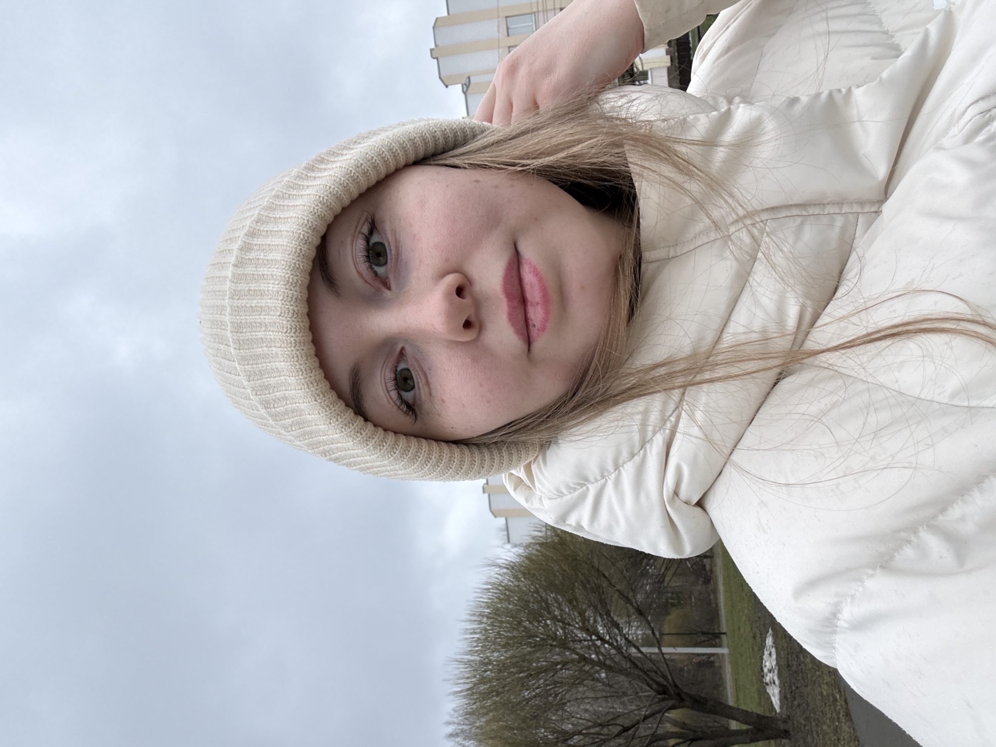 A young woman, likely in her late teens or early twenties, is featured in a close-up selfie, oriented as if the image has been rotated 90 degrees counter-clockwise. She has fair skin, light brown or blonde hair that falls over her shoulders, and striking green eyes. She looks directly at the camera with a calm, neutral expression, her lips slightly parted and colored with light pink lipstick.
She is dressed in a light beige or off-white knit beanie that covers her ears and forehead, and a matching or similarly colored quilted puffer jacket, indicative of cool weather. Her right hand is visible near the top of her head, possibly adjusting her hair or beanie as she holds the camera. Tiny water droplets are visible on the fabric of her jacket, suggesting light precipitation or mist.
The setting is outdoors, under an overcast, light grey sky that dominates the upper portion of the background. To her left, a multi-story, light-colored apartment building stands. Below the building and partially obscured by her, bare trees with intricate branches are visible, suggesting late winter or early spring. A patch of green grass is also discernible. The overall impression is a daytime scene in an urban or suburban environment. There is no visible text in the image.