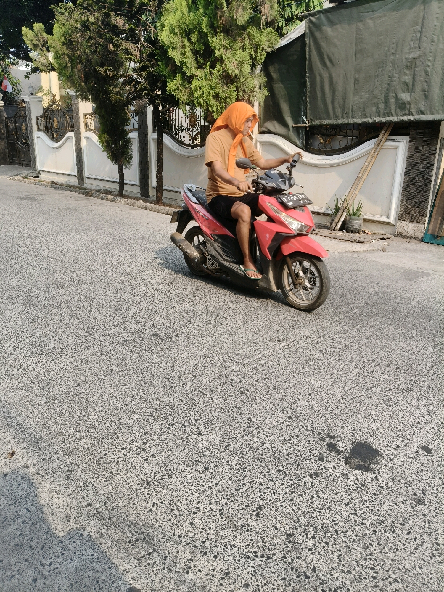 A medium shot captures a person riding a red scooter on an asphalt road under a sunny sky. The individual, wearing an orange head covering and an orange t-shirt, is seated on the scooter, which has a license plate that reads "B 4796 TRP". The background features a white fence with black ornate gates, trees with green foliage, and a building covered with a dark green tarp. The scene appears to be in a residential area in South Tangerang, Indonesia.