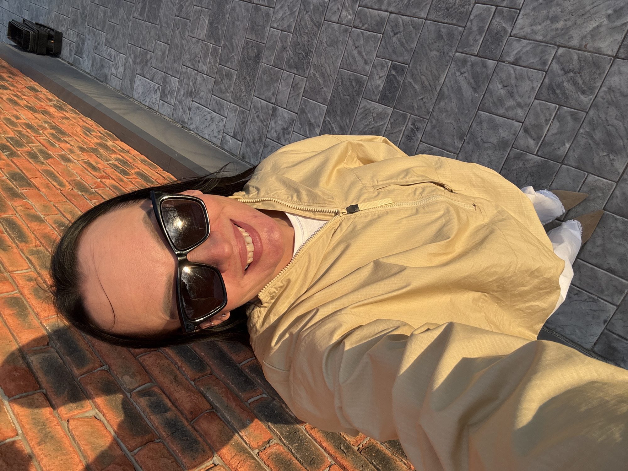 The image shows a woman lying on a brick sidewalk in Chaikovsky, Russia. She is wearing sunglasses and a tan jacket and is smiling. She is wearing white pants and tan shoes. Next to the sidewalk is a gray brick wall. There is a black object located along the wall.