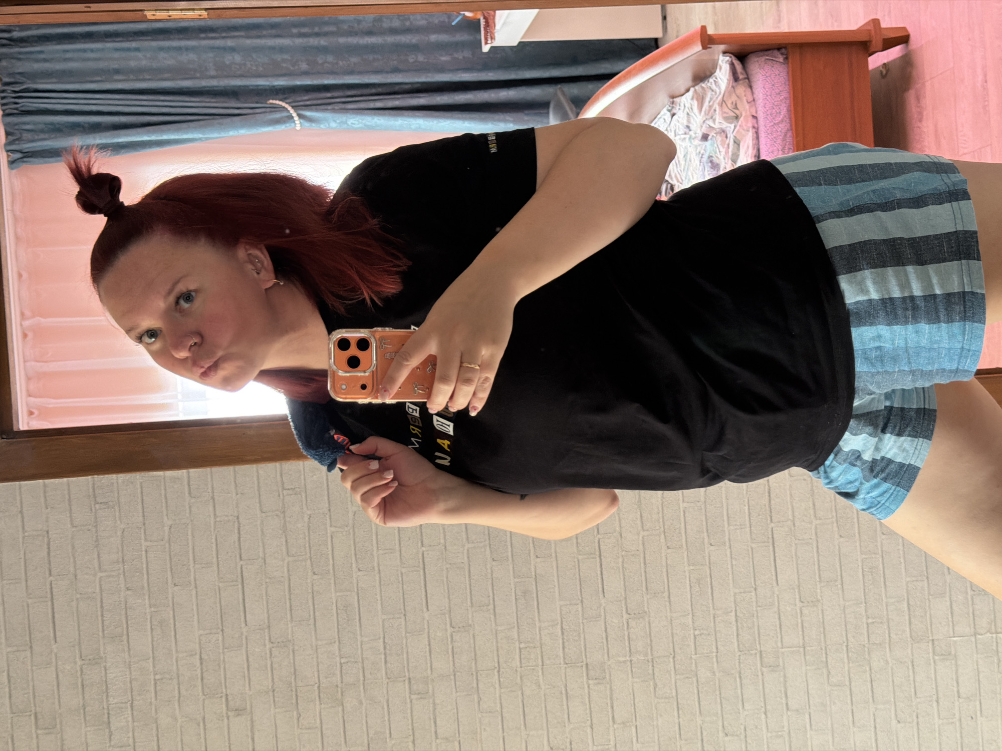 A woman is taking a selfie in what appears to be a room of a house. The woman has fair skin, red hair styled in a bun on top of her head, and is wearing a black t-shirt and blue striped shorts. She is holding an orange phone in her right hand.
The background of the image shows elements of the room. There is a blue curtain visible near the top left of the image, a bed with a pink blanket and pink pillow behind a bed frame, and a brick wall is visible behind the woman on the bottom of the image.
The location of the city of Chaikovsky, Russia cannot be confirmed in this image.