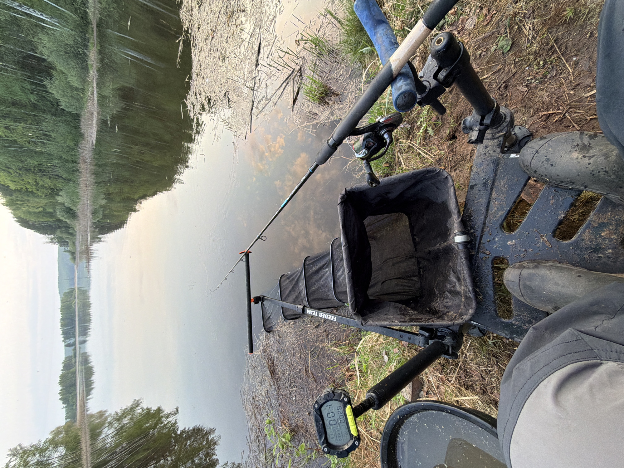A scene at a body of water, likely a lake or a wide river, adjacent to a grassy and wooded area in Chaykovsky, Russia. A fishing setup is present, consisting of a fishing rod with a reel, a black mesh net, and a black support stand. An electronic device, possibly a timer, is attached to the fishing setup. An individual wearing grey pants and rubber boots is also visible. The water's surface has debris floating on it.