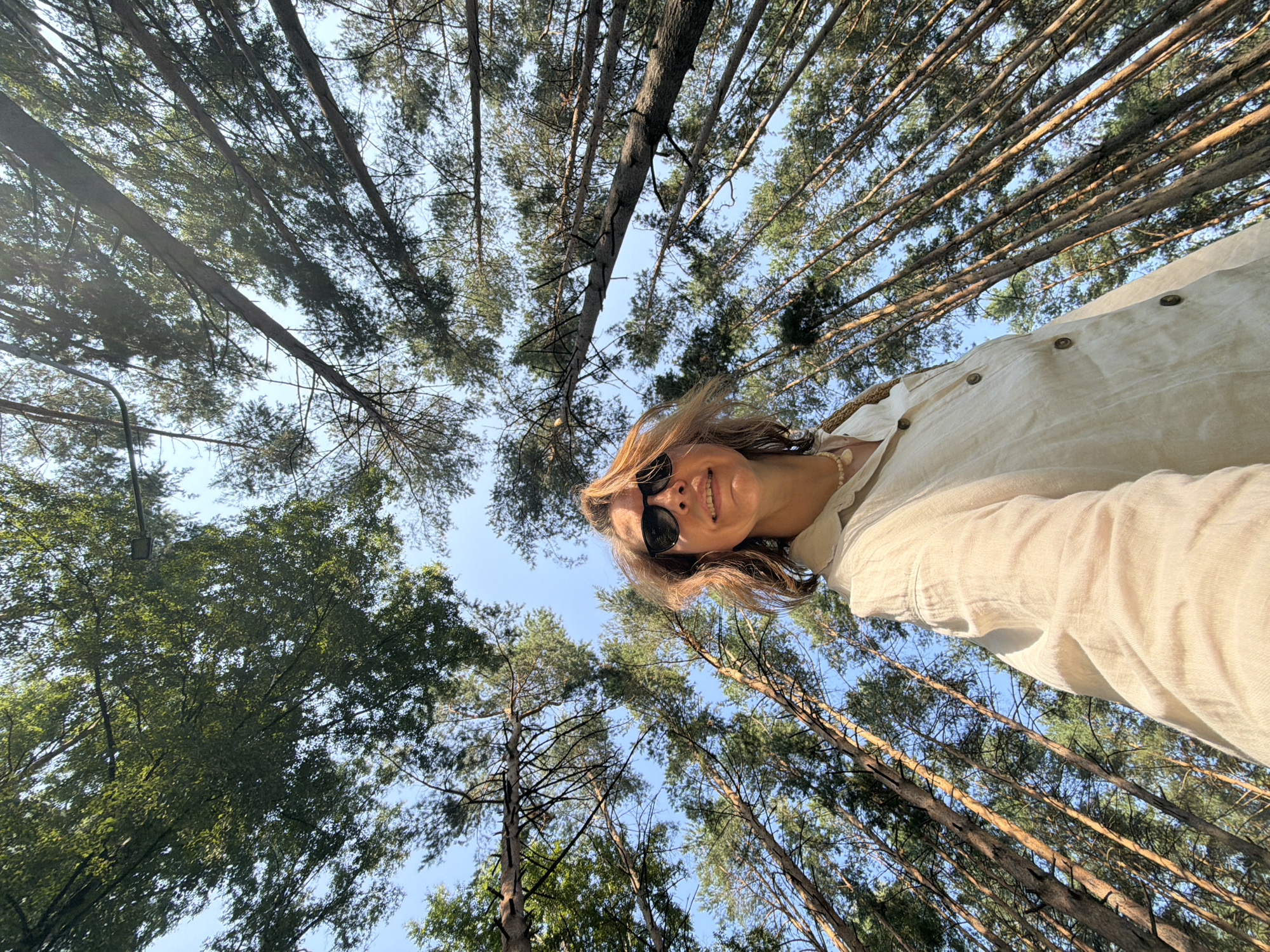 In the city of Chaikovsky, Russia, a low-angle shot captures a woman with brown hair and sunglasses looking up and smiling. She is wearing a light-colored linen shirt and a beaded necklace. The background is a dense forest of tall pine trees, with their branches extending towards a clear blue sky. The sun is casting light through the canopy, creating a dappled effect on the trees and the woman. The overall impression is one of a sunny day spent outdoors in nature.