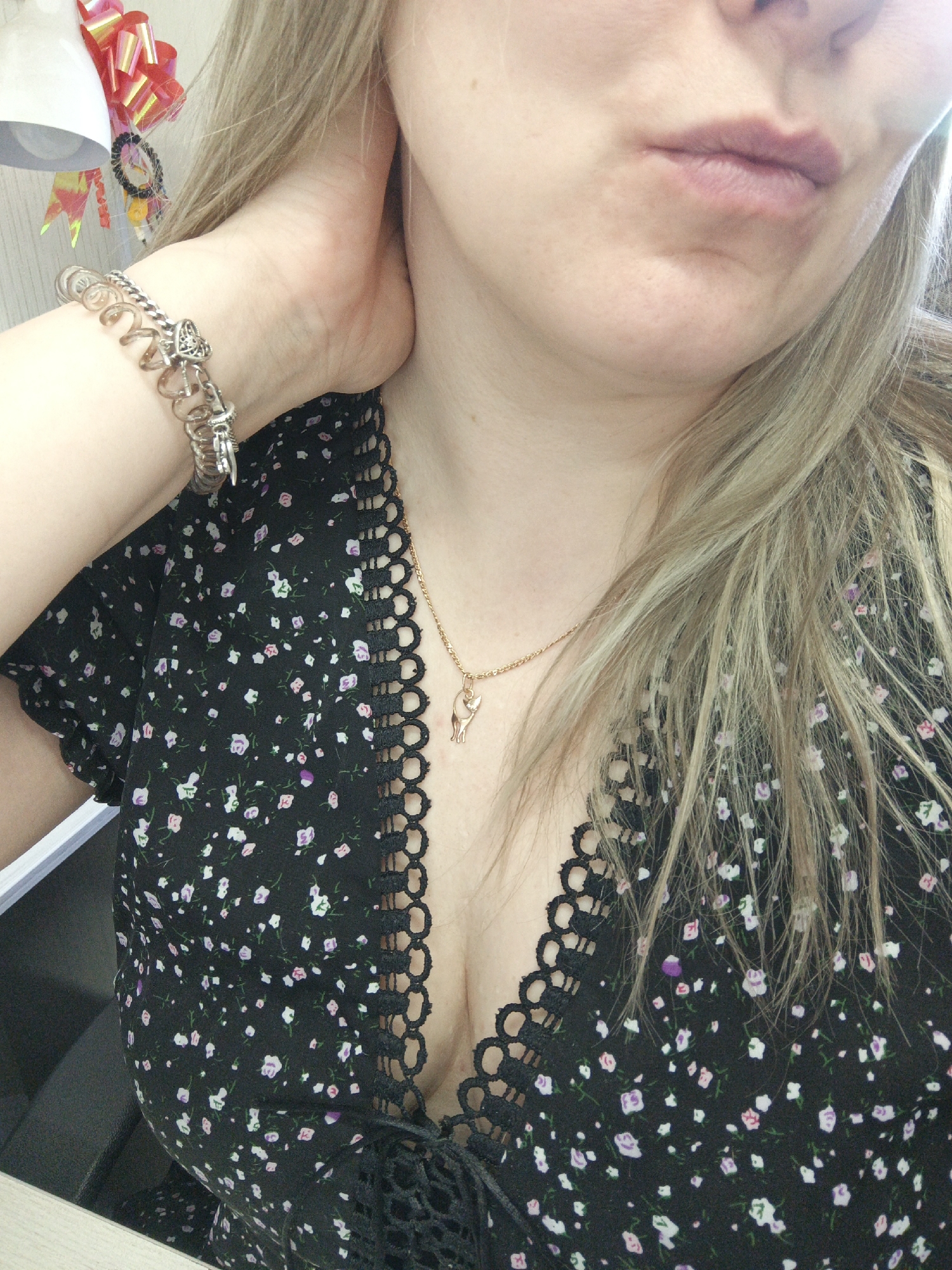 A selfie shows a woman with blonde hair, pale skin, and visible lips. She wears a black floral dress with a lacy trim and a gold necklace with a cat pendant. On her wrist is a silver charm bracelet. Her hand is touching her neck. The background includes a desk, an office chair, and some decorations. There is no verifiable evidence to confirm the image was taken in the city of Noviy, Russia.