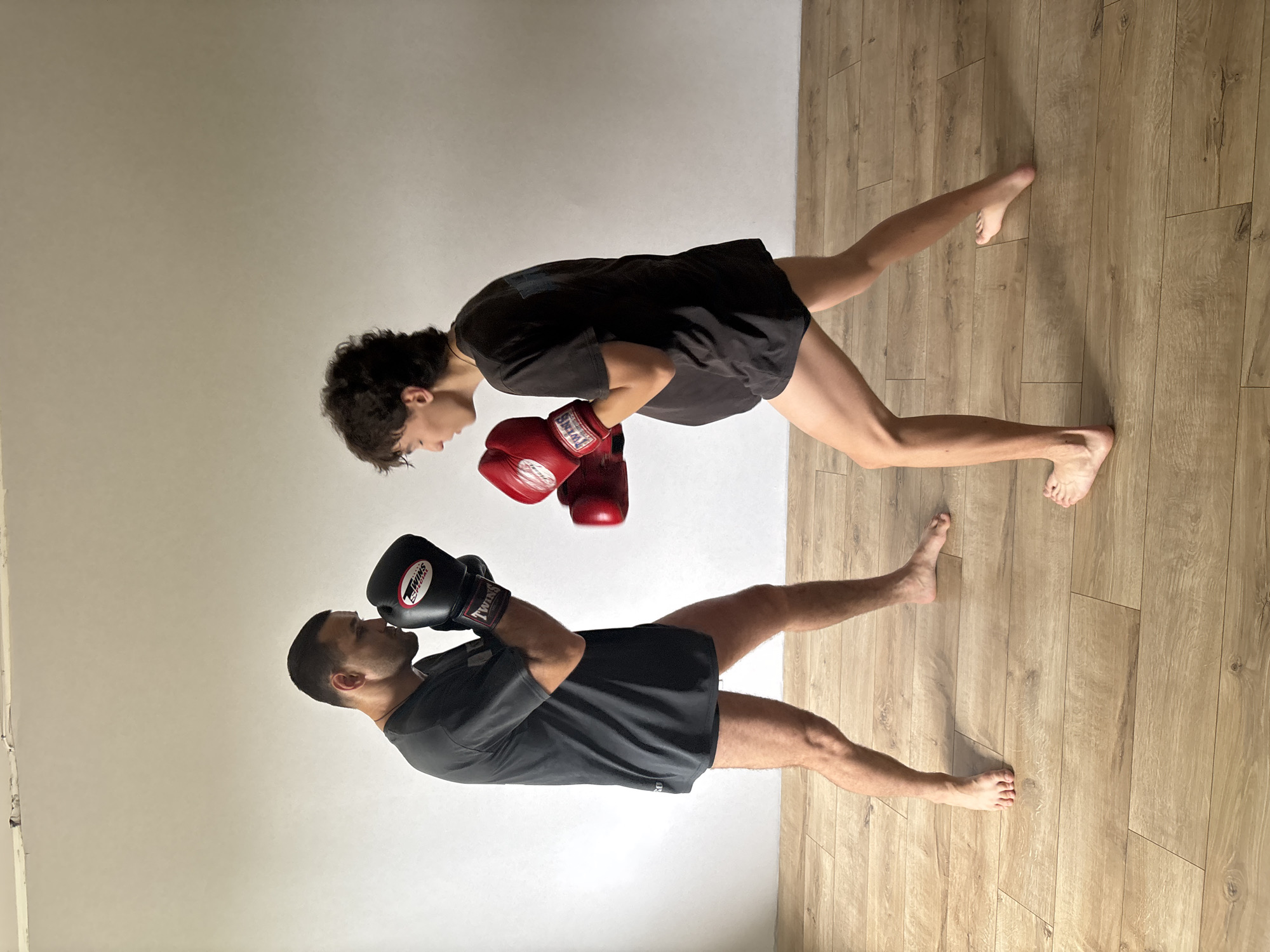 The image displays two individuals, likely engaged in a boxing or martial arts training session, indoors in Chaikovsky, Russia. The plain, light-colored wall serves as a backdrop, and the floor is covered with light brown wooden planks.
On the left, a man with short dark hair and a beard is depicted in a fighting stance. He wears a dark grey t-shirt, dark shorts, and black boxing gloves, which clearly show the brand "TWINS". He is barefoot and looks upwards, directing his gaze towards the other person.
Opposite him, on the right, another person with curly dark hair is also in a similar stance. They wear a black t-shirt, dark shorts, and red "TWINS" boxing gloves. This individual is also barefoot and looks downwards, facing the man. Both participants have their gloved hands raised, suggesting readiness for combat or a posed moment during training. Their expressions appear focused and serious.