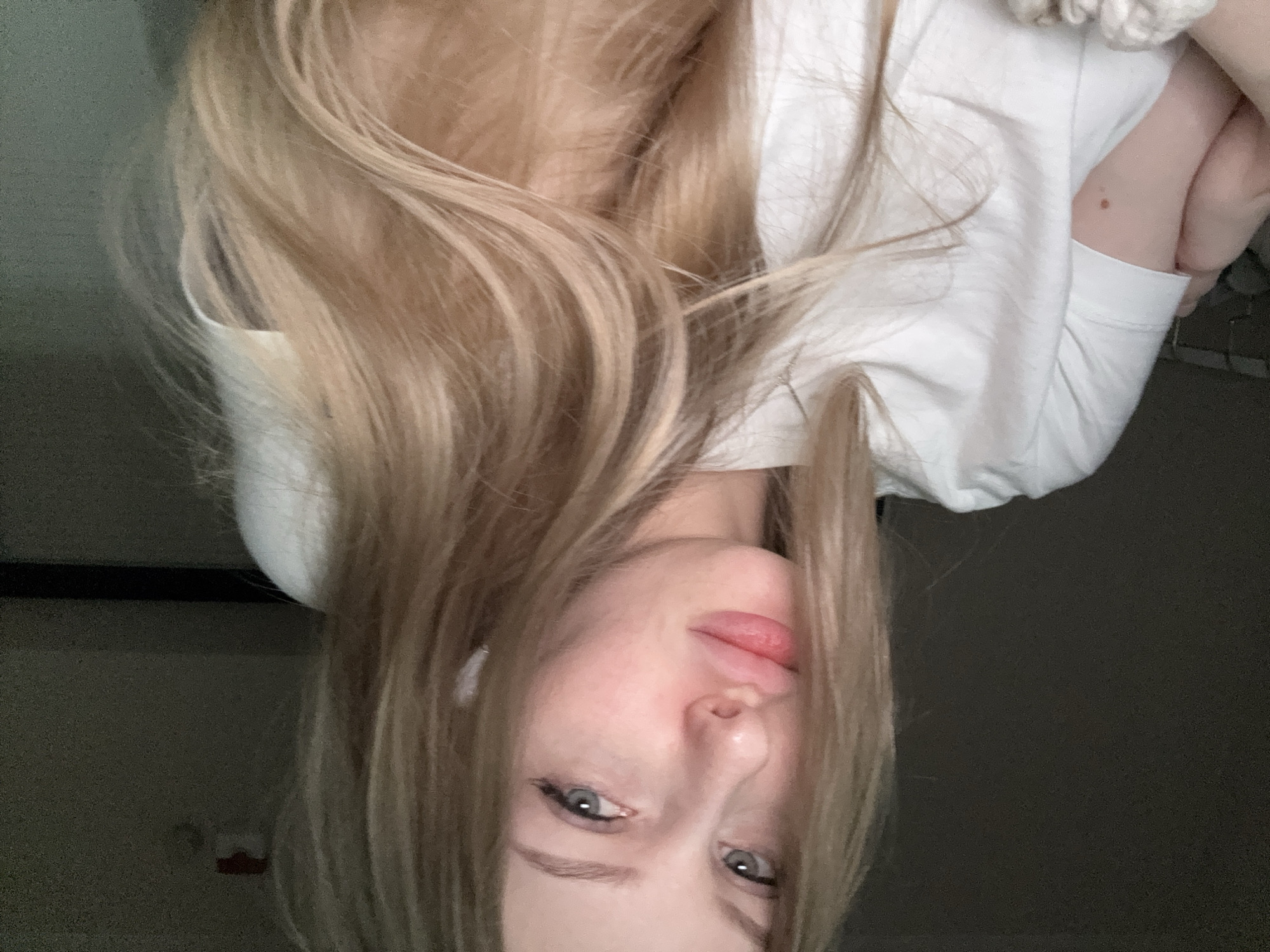 The image is a portrait, rotated 90 degrees clockwise, featuring a young individual, likely female, with light blonde hair. The subject's head and upper body are visible, with the long, straight hair fanned out around them. Their eyes are light blue or grey, directed towards the camera with a neutral expression. Subtle eyeliner is visible on the upper lash line. The lips are naturally colored and appear slightly parted.
The individual is wearing a plain white long-sleeved t-shirt or sweatshirt. A white, soft, textured object, possibly a scrunchie, is visible near the upper right shoulder. A small, brown mole or birthmark is visible on the inner arm.
The background is dark and indistinct, suggesting an indoor setting with low or indirect lighting. No specific architectural features or objects are clearly identifiable, though there's a hint of a dark surface on the left and a darker, textured area on the right. There is no visible text in the image.