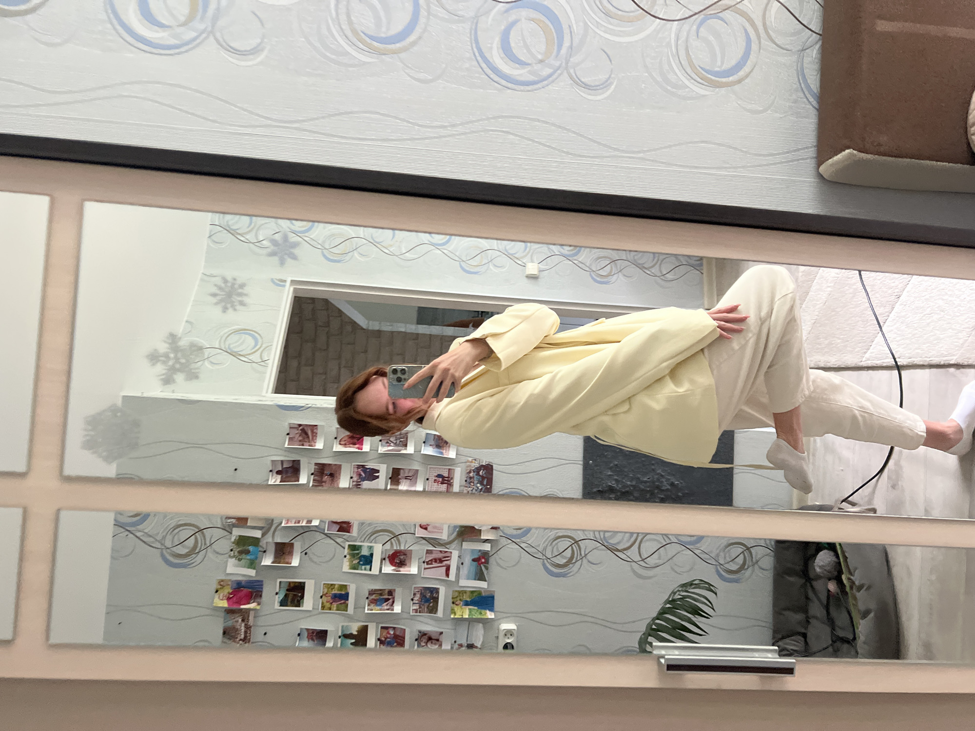 The image captures a mirror selfie of a young woman inside a residence located in the city of Chaikovsky, Russia. The woman, appearing to be in her 20s to 30s with reddish-brown hair, is reflected in a multi-panel mirror, likely part of a wardrobe or full-length wall unit, framed in a light brown material. She is dressed in a light pastel yellow oversized blazer and light cream-colored trousers, with white no-show socks. Her right leg is bent at the knee, heel lifted, as she holds a grey smartphone with a triple-lens camera system in her right hand to take the photograph. Her fingernails exhibit light red or pink polish.
The reflected background features light-colored wallpaper adorned with blue, brown, and grey swirling patterns, interspersed with abstract snowflake designs. On the left wall, numerous small, square, polaroid-style photographs depicting various individuals and scenes are pinned, forming a dense collage around a visible white electrical outlet. A dark grey upholstered pet bed rests on the floor to the left, positioned near a green leafy plant. A black electrical cord traverses the light-colored, textured floor, likely carpet. To the upper right of the reflection, a doorway opens into another room with a light-colored brick wall. A portion of a light brown upholstered sofa or chair is visible in the far right reflection.